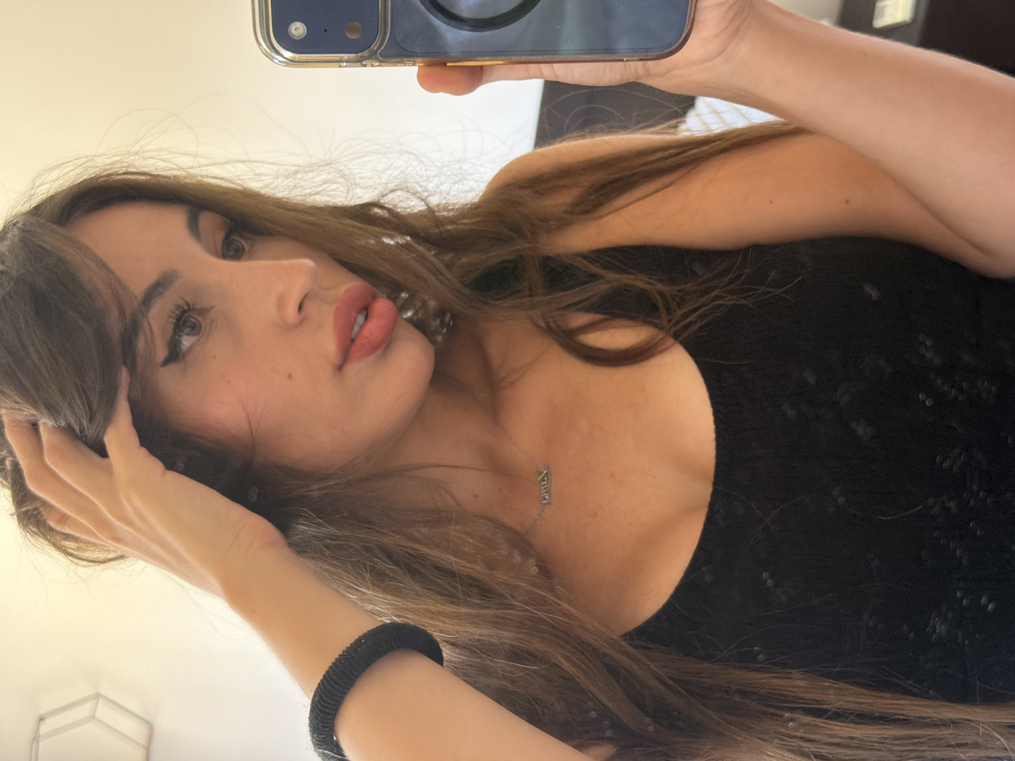A young woman with long, brown hair, wearing a black top, is taking a selfie in a room. She is holding her hair up with her right hand and her left arm is bent at the elbow, with a black bracelet on her wrist. Her head is tilted to the right, and she is looking to the side. The room appears to be a bedroom, with a bed and a dresser visible in the background. The lighting is soft and natural.