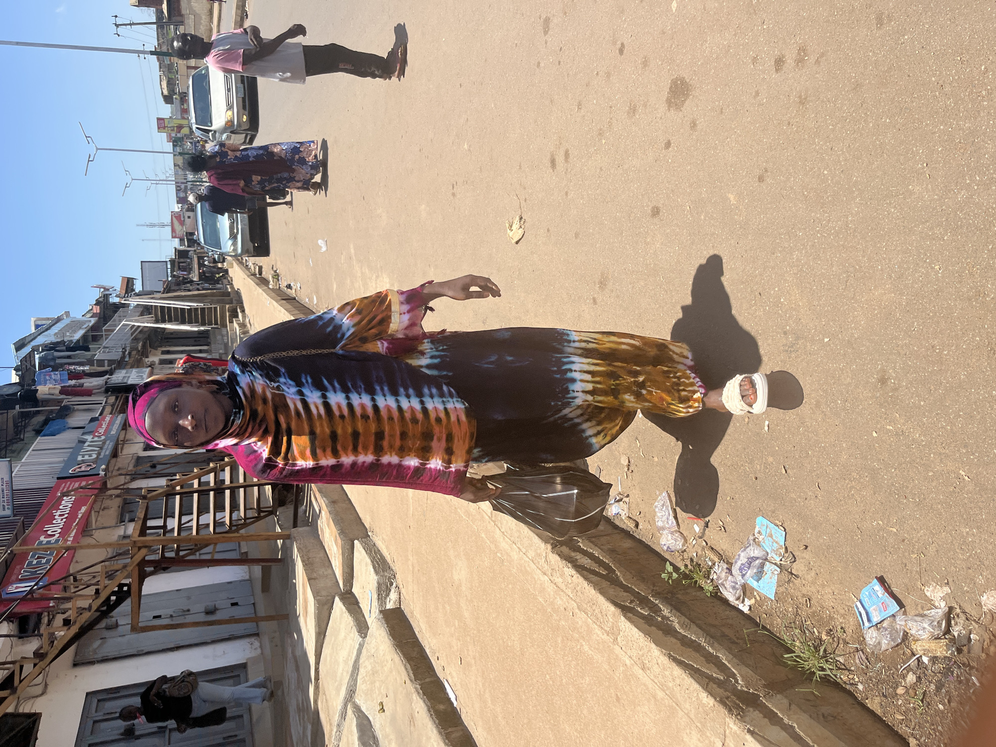 A medium shot shows a woman in a vibrantly colored tie-dye dress and headscarf walking on a paved street in Jos, Nigeria. The woman is in the foreground, slightly off-center to the right, and she is looking directly at the camera. She carries a black plastic bag in her left hand. Her right arm is relaxed by her side. She is wearing white slide sandals.
Behind her, the street continues, with other pedestrians and vehicles visible in the middle ground. A silver sedan and a light-colored SUV are present on the road. The woman is walking on the right side of the street, and there is a raised concrete sidewalk to her left. On the sidewalk, there are stairs leading up to buildings and shops. Various signs are visible on the buildings, including "KEZ Collections" and "ELVTE Collections". Litter, including plastic bags and wrappers, is scattered on the sidewalk and on the street. The sky is clear and blue. The scene appears to be a commercial area on a sunny day.