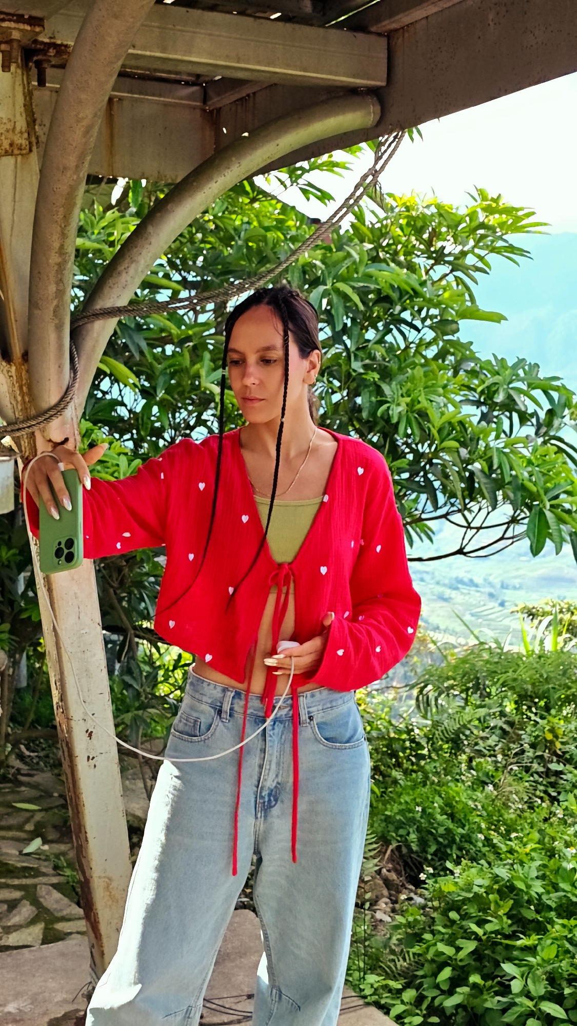 An outdoor portrait in Tu Le, Vietnam, features a young adult female facing slightly left, holding a smartphone and a charging device. She has dark hair styled in braids and wears a red, long-sleeved, tie-front crop top adorned with white heart patterns, a light green bralette underneath, and light blue denim jeans. In her left hand, she holds a green-cased smartphone (likely an iPhone) pointed towards herself, consistent with taking a selfie or video. A white charging cable extends from the phone to her right hand, which holds a small white object, possibly a power bank or charging adapter. A rusty metal pole with a rope wrapped around it is visible on the left side of the frame. The background consists of dense green foliage and trees, with a clear blue sky and distant mountainous landscape, characteristic of terraced rice fields, under bright daylight.
