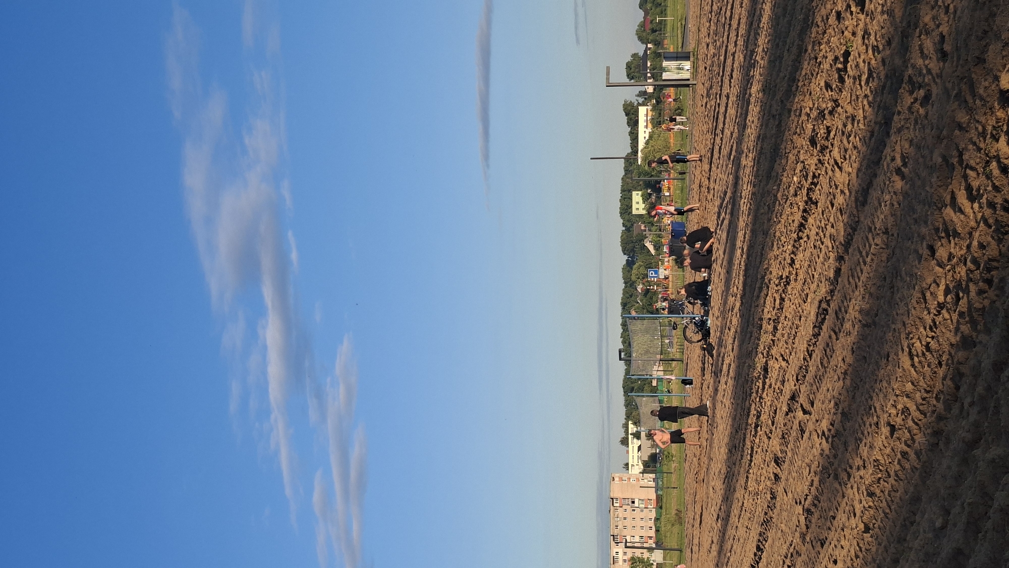 The image depicts an outdoor recreational area in Smyavichy, Belarus, characterized by a sandy ground, likely a beach or sports field. In the foreground, the sand shows tire tracks, indicating recent vehicular or equipment use. A group of individuals are visible on the sand. Some are standing near a volleyball net structure, while others are seated. A bicycle is leaning near the net. In the middle ground, there are more people, some standing and engaged in activity, others seated. A volleyball net is prominently positioned. Beyond the sandy area, a fence separates it from a green space with trees. In the background, there are buildings, including multi-story apartment blocks and smaller structures, suggesting an urban or suburban setting. The sky is a clear blue with scattered white clouds, indicative of daytime.