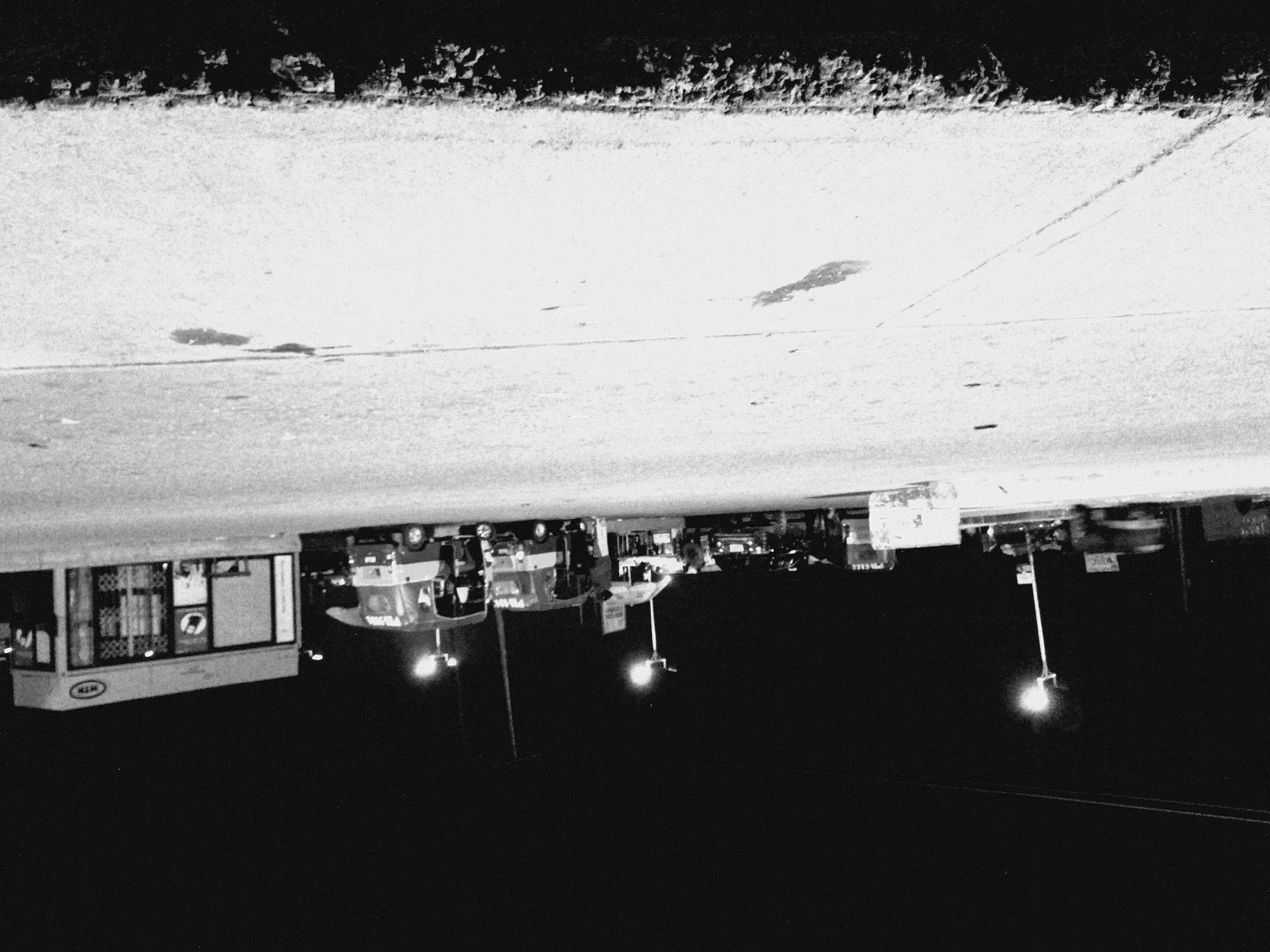 In Bukuru, Nigeria, a nighttime scene unfolds in front of a commercial building with a "MTN" logo. Several vehicles are parked on a paved surface, including what appear to be three distinct auto-rickshaws or similar three-wheeled passenger vehicles and two cars. One car, positioned centrally, displays what might be police markings. An auto-rickshaw on the left has a license plate that partially reads "PB 716". Two individuals are visible standing near one of the auto-rickshaws. Streetlights illuminate the area, casting a stark, high-contrast ambiance. The foreground shows a rough, textured surface.