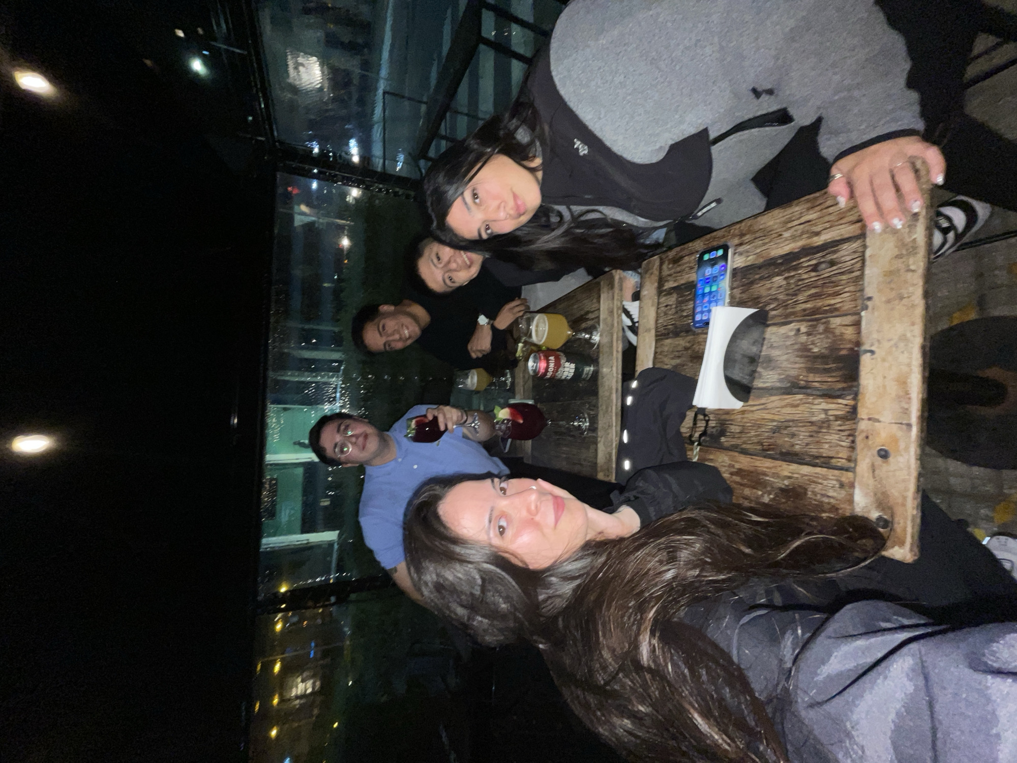 At night, a group of six individuals are gathered around a wooden table, likely in a restaurant or bar in Buenos Aires, Argentina, given the urban cityscape visible through the window. The composition includes: four men, two women. The individuals are smiling and appearing to interact in a social setting. Beverages, in glasses and cans, are on the table, as well as a cell phone. The environment includes a view of city lights visible through a window behind the group.