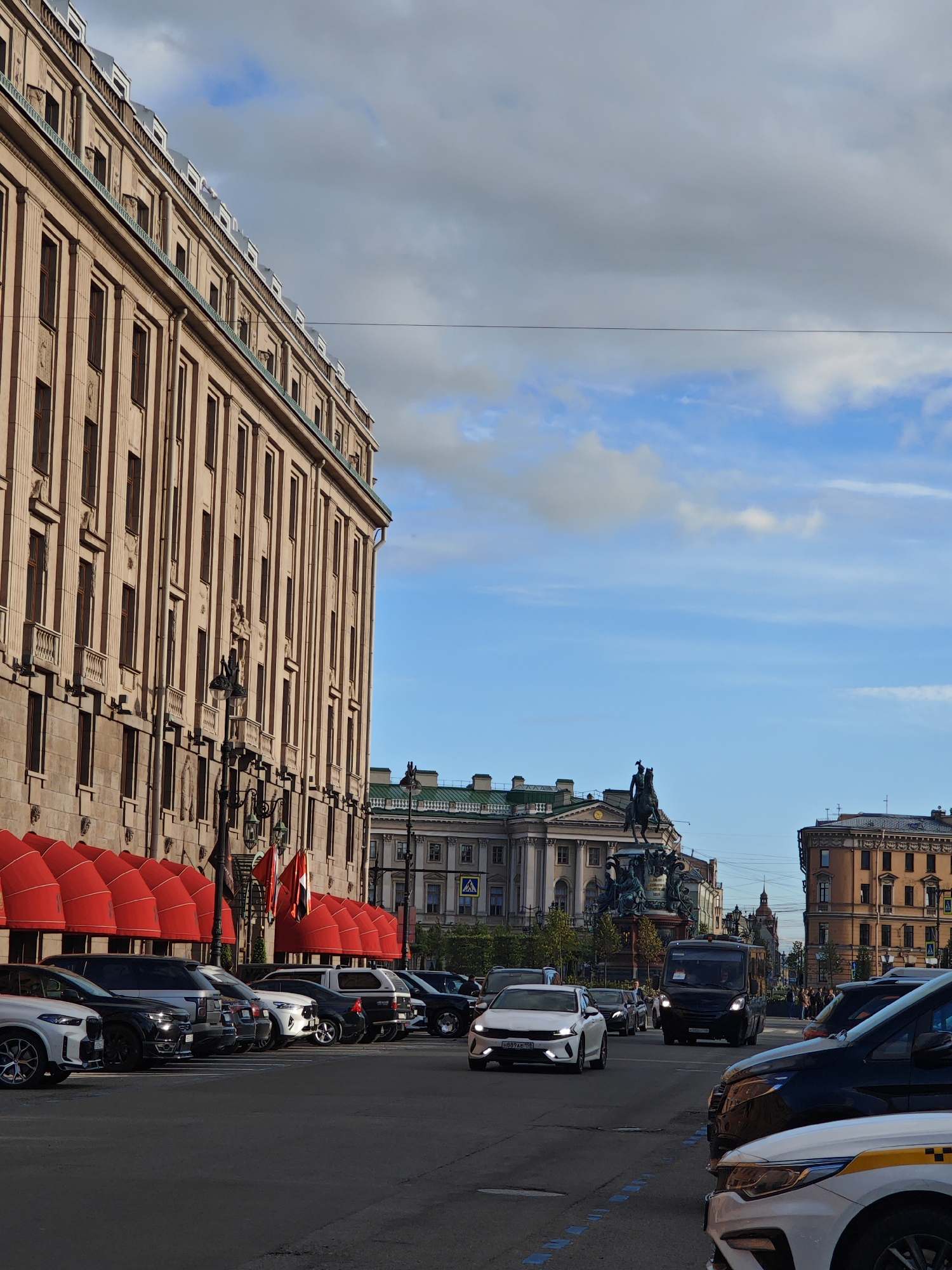 The image depicts a street scene in Saint Petersburg, Russia, likely near Palace Square. On the left, a grand building with a light brown facade and numerous windows dominates the foreground. Red awnings are visible along the ground floor. In the middle ground, a bronze equestrian statue stands atop a monument, silhouetted against a bright blue sky with scattered clouds. Several cars, including a black van and a white car, are driving on the street. The overall atmosphere suggests a bustling urban environment with historical architecture.