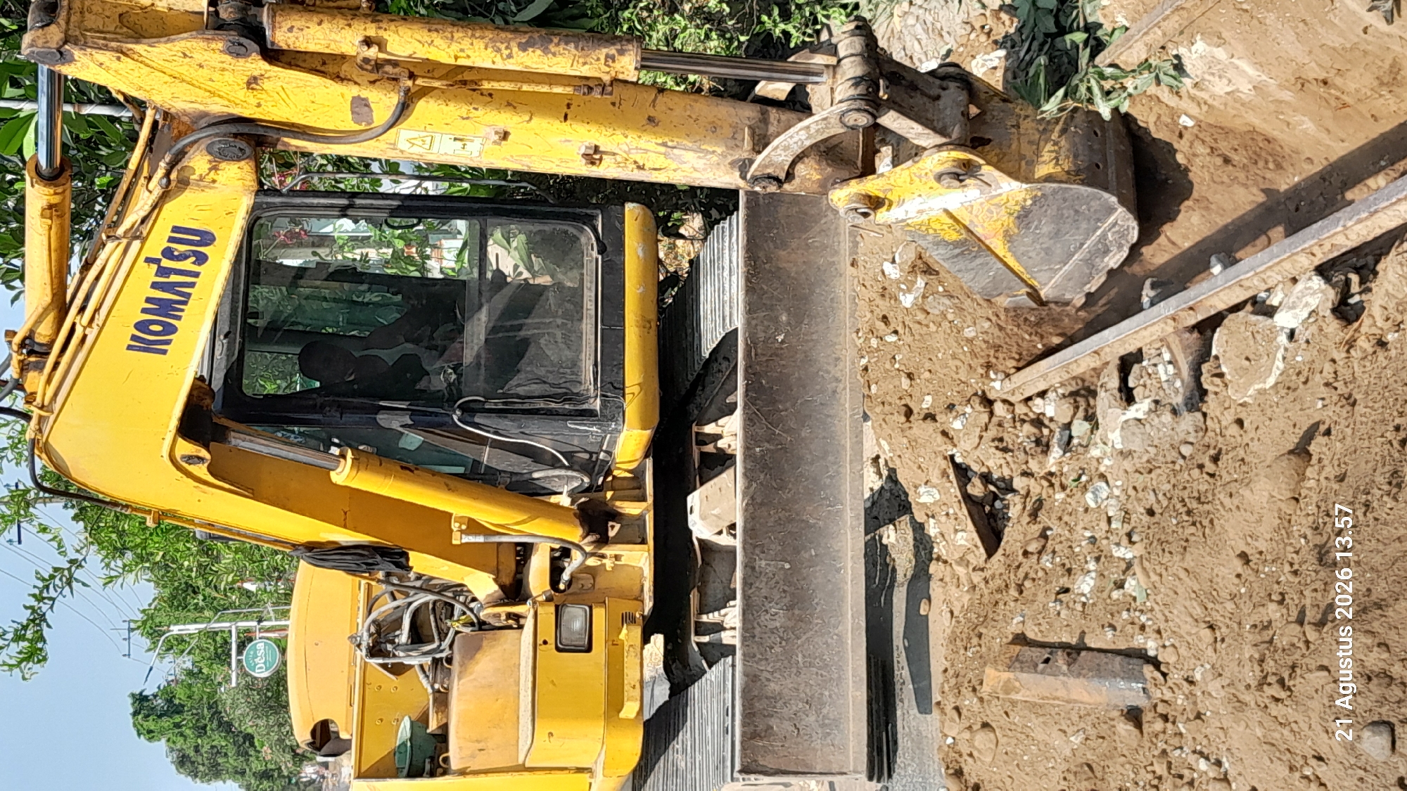 An excavator, identified by the "KOMATSU" logo, is actively digging a trench on a dirt road. The machine is yellow and its tracks are visible. The cabin of the excavator shows a silhouetted figure, presumably the operator. The surrounding environment consists of loose soil, rocks, and some green foliage. A timestamp in the bottom left corner indicates "21 Agustus 2026 13.57," suggesting the image was taken on August 21, 2026, at 1:57 PM. The scene appears to be in Takeran, Indonesia, given the context of the question.