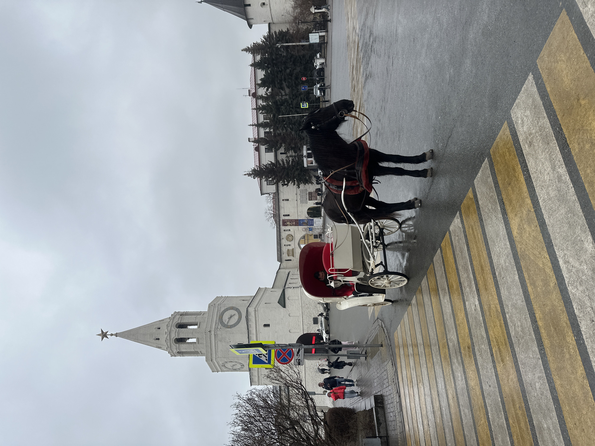 A horse-drawn carriage is on a crosswalk in Kazan, Russia. The horse is black, and the carriage is red and white. A tall, white stone building with a clock and a star at its peak is in the background. Several people are walking on the sidewalk near the building. The weather appears overcast.