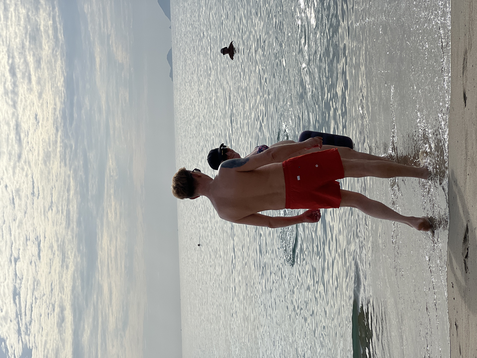 In Nha Trang, Vietnam, the scene depicts a calm ocean with a sandy shore under a cloudy sky. In the foreground, a shirtless man wearing red swim trunks stands with his back to the camera, facing the water. To his left, a woman wearing a bikini and a black baseball cap is partially visible, also facing the water. She appears to be holding onto something or someone behind her. Further out in the ocean, another individual is swimming, with only their head visible above the water. In the distance, islands can be seen on the horizon. The water has gentle ripples, and a slight wave is washing up onto the sand where the man is standing. The overall atmosphere is relaxed and summery.