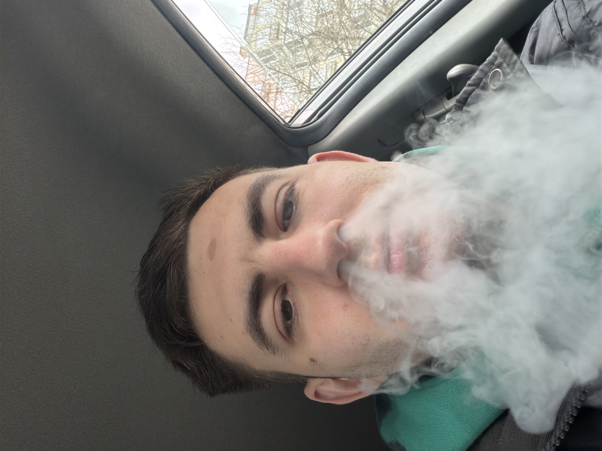 A close-up photograph, rotated counter-clockwise, depicts a Caucasian male exhaling a large cloud of vapor. The man has short brown hair, brown eyes, and fair skin with a mole on his forehead. He is positioned inside a vehicle, likely a car, as indicated by the grey interior and sunroof visible at the top of the frame, which offers a view of bare tree branches and a building exterior. A green fabric and part of a grey jacket can be seen beneath the vapor cloud. The precise location in Varna, Bulgaria, is undeterminable from the visual data provided.