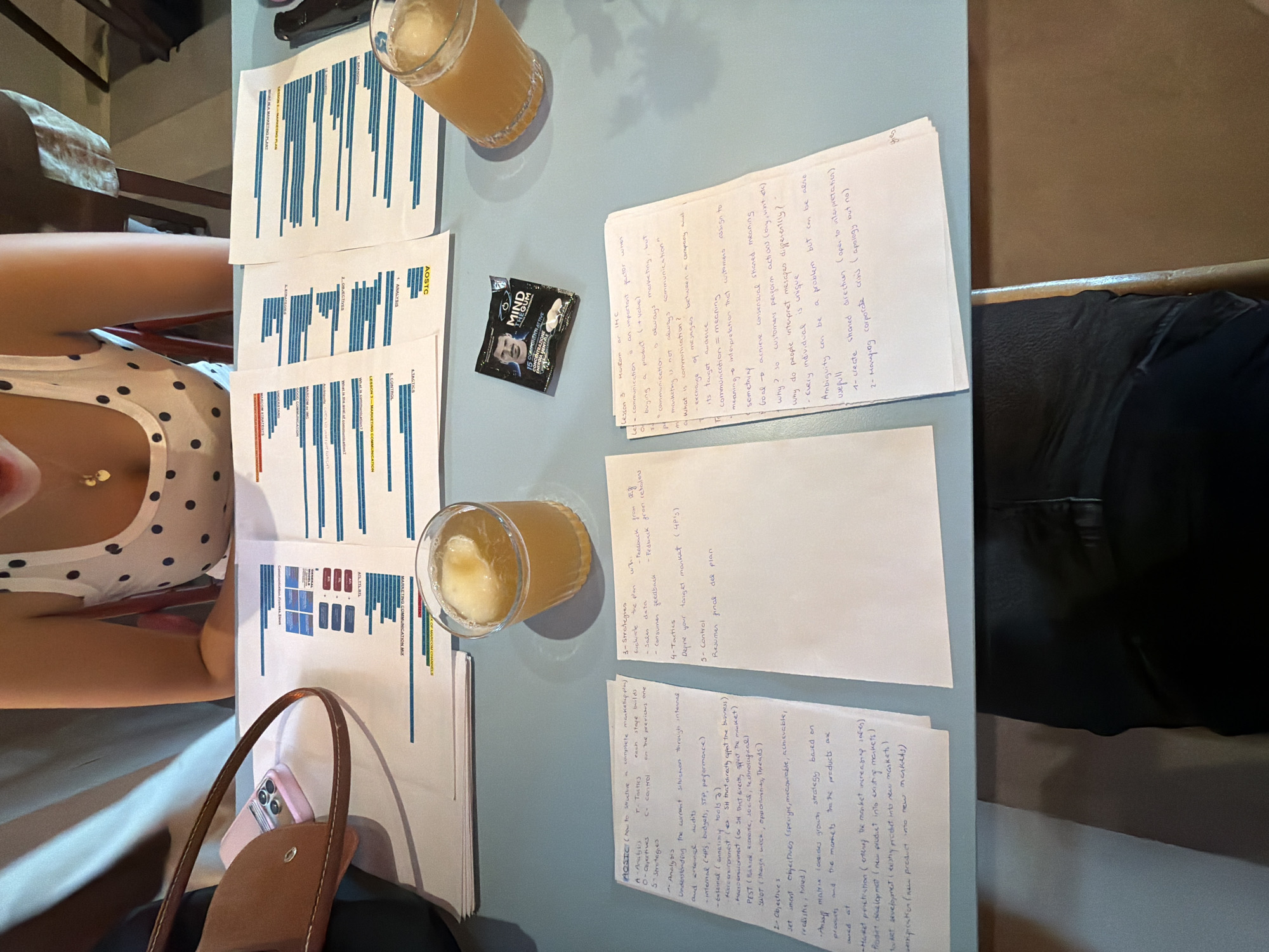 An overhead, medium shot captures a section of a light blue table, likely in an office or cafe setting, with a partially visible person sitting at it. The person, only seen from the chest up, wears a white tank top with black polka dots and a delicate gold necklace. Spread across the table are several sheets of paper covered in handwritten notes and printed charts. To the left, a brown purse and a pink smartphone are visible. In the center of the table, a pack of "MIND GUM" is placed next to a glass containing an amber-colored beverage with ice. The lighting suggests an indoor environment, possibly during the day, with a soft shadow cast by the person and objects. The content of the papers appears to be business-related, with headings like "AOSTC," "Lesson 3-Marketing Communication," and diagrams featuring bar charts. The overall scene conveys a sense of work or study in progress.