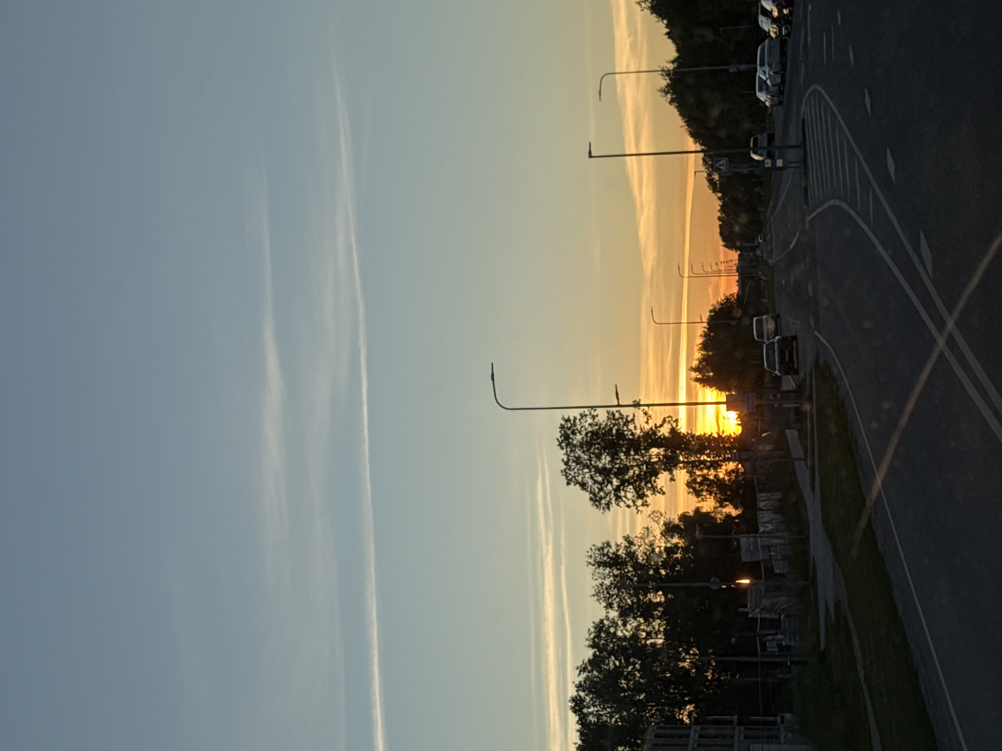 A street scene in Tallinn, Estonia, during sunset. The sky displays a gradient of blue to orange, with thin, elongated clouds. Trees line the right side of the road, silhouetted against the bright sky. Several streetlights are visible along the road. On the left, there is an incomplete building structure and a fence. The road itself is paved, with white lane markings. Several cars are parked on the right side of the road and some are driving on the road. One car is visible on the left side of the road.