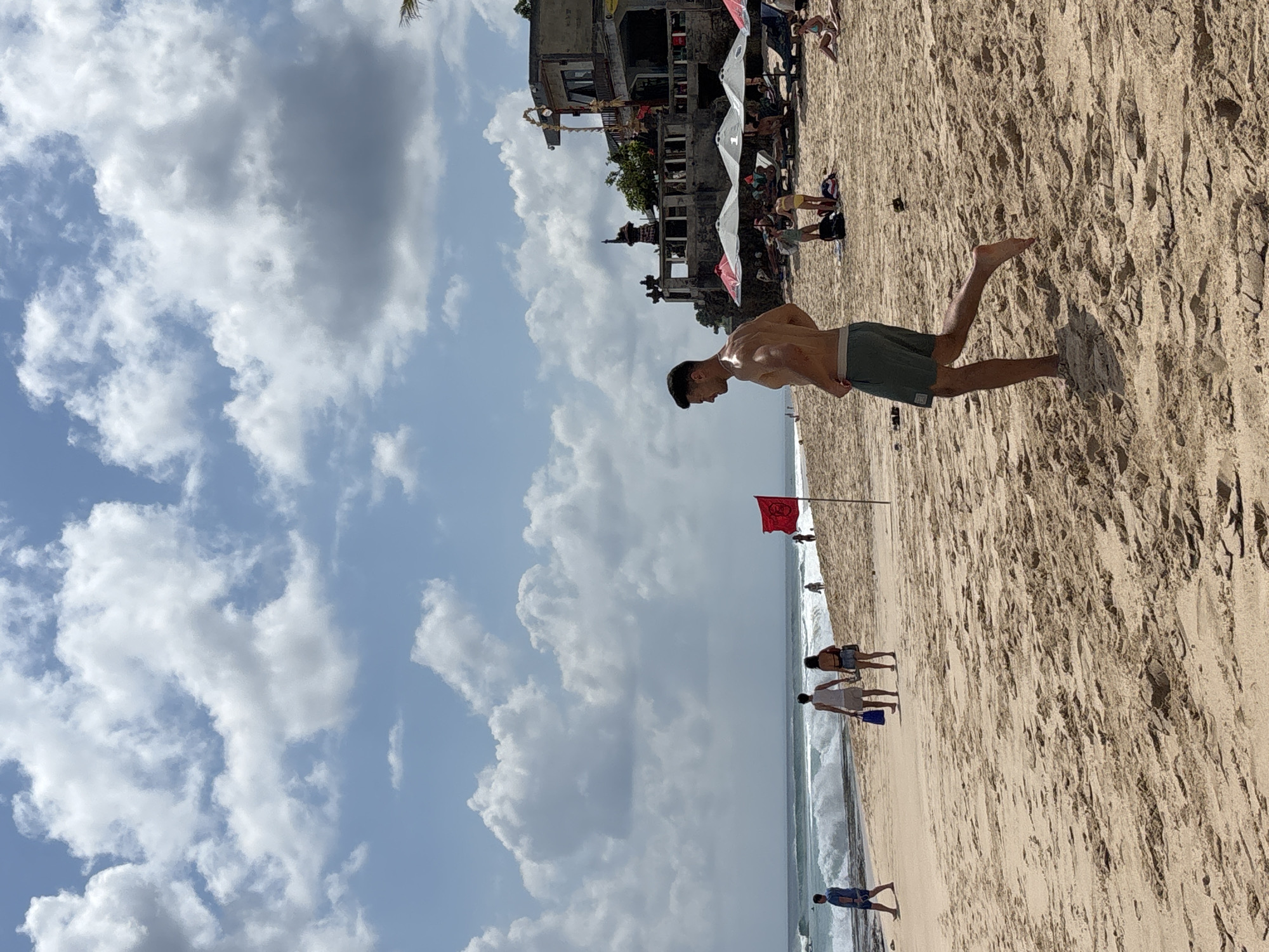 The image depicts a sunny day at a beach in Kuta Selatan, Indonesia. In the foreground, a shirtless man with dark hair is running on the sandy beach. He is wearing dark green shorts. In the mid-ground, several people are visible. To the left, a couple walks away from the camera. Further down the beach, a red flag is planted in the sand. Behind the flag, there are more beachgoers and some beach umbrellas. In the background, the ocean with waves is visible, and a multi-story building with a stone facade and balconies stands on a rocky outcrop. The sky is blue with scattered white clouds.
