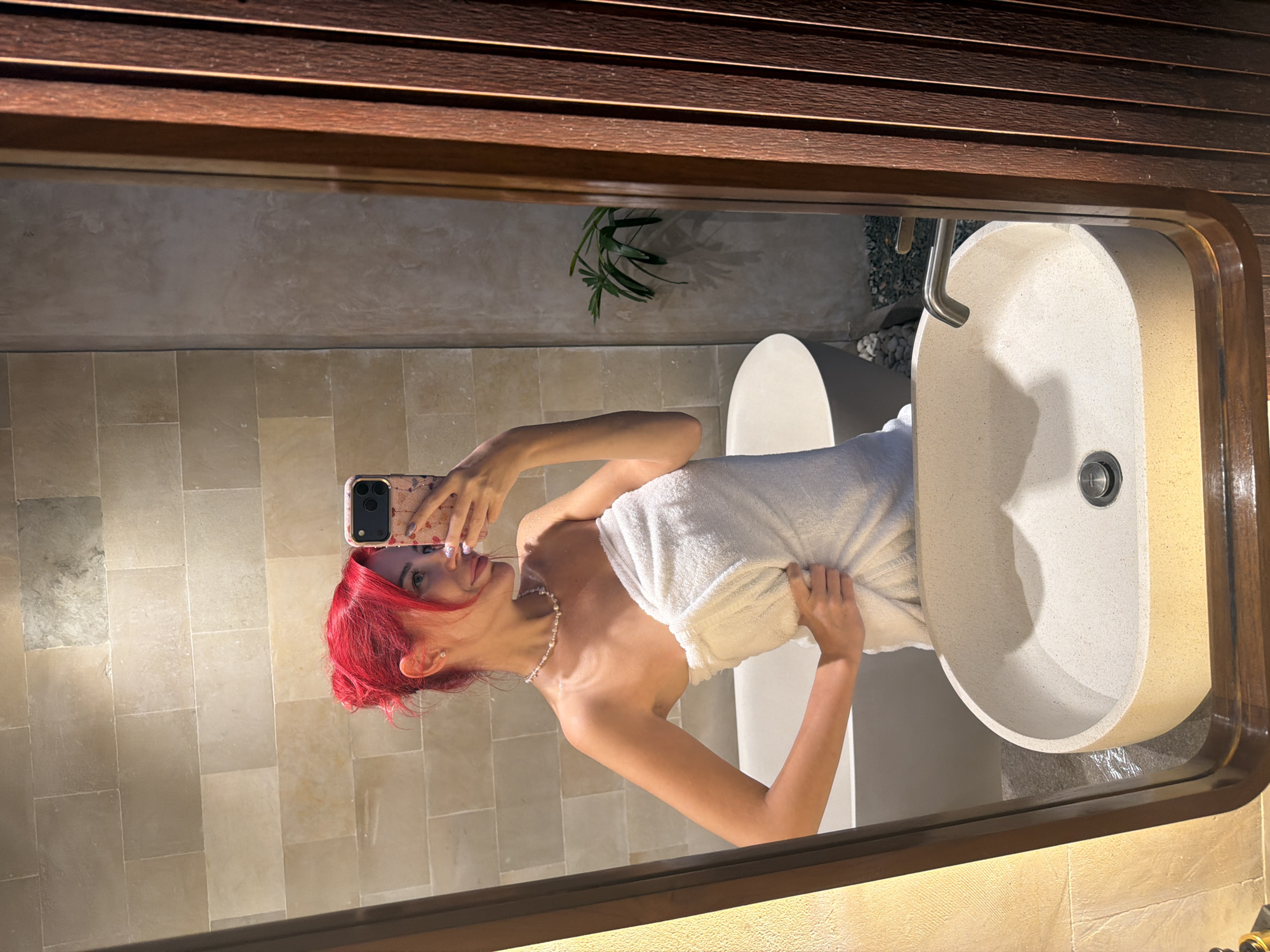 A young woman with bright red hair, wearing a white towel and a pearl necklace, takes a selfie in a bathroom mirror. The bathroom has a modern design with beige stone tiles on the walls and a speckled oval sink. A bathtub and a small plant are visible in the reflection. The woman is holding her phone, which has a patterned case, and is smiling. The image appears to be taken in Kuta, Indonesia.