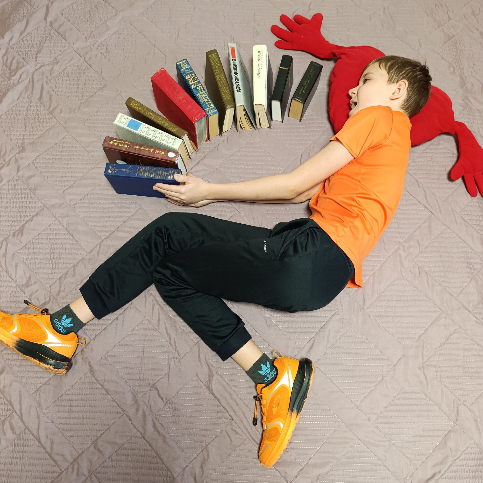 A boy, dressed in an orange t-shirt and black pants, lies on a bed with a red pillow shaped like a person. He has a row of books balanced on his hands and arms. The books appear to be in Russian, including works by Dostoyevsky, Bulgakov, and Pasternak. The boy is wearing orange sneakers with black socks. Based on the books and location (implied from text recognition), this is likely in a Russian-speaking region.