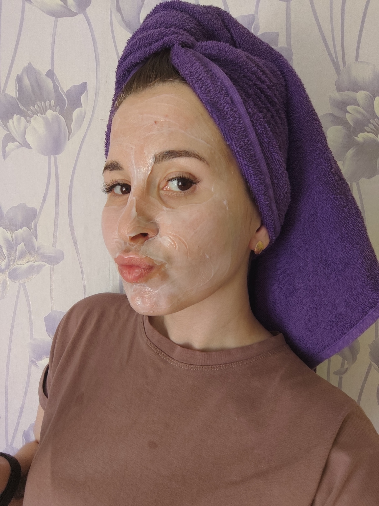 A young woman is posing for a photo while wearing a face mask and a purple towel wrapped around her head. She is wearing a brown t-shirt. The background is a wallpaper with a floral pattern. This location is not in Фоки, Russia.