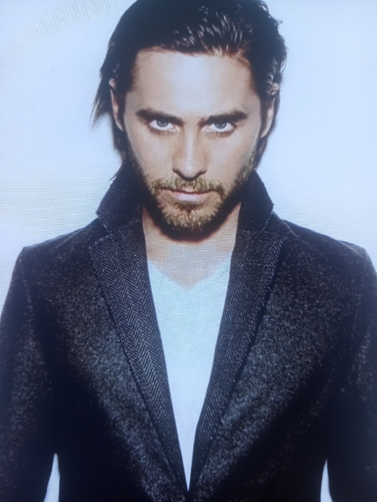 This media file is a portrait of a man. He has dark, medium-length hair that is swept back from his face, and piercing blue eyes. He has a light beard and mustache. He is wearing a dark, textured jacket with a herringbone pattern and a lighter-colored shirt underneath. The background is plain white, creating a studio-like setting. The lighting is bright and even, with no shadows indicating a specific time of day or weather conditions. The man's expression is intense and direct, looking straight at the camera. The image appears to be a posed photograph, likely for a magazine or promotional material, given the clean and stylized presentation. There is no visible text in the image.