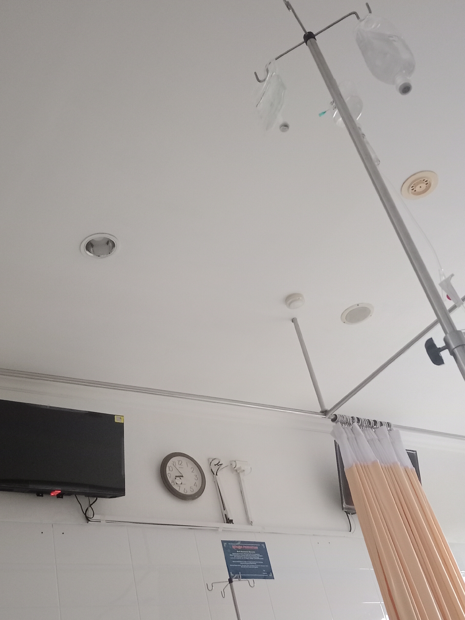 A hospital room in Yogyakarta, Indonesia, features an IV pole with multiple bags of fluid hanging from it, a black television mounted on a white tiled wall, and a round wall clock. A pale orange curtain is partially drawn, hanging from a metal rod. The ceiling has recessed lights and a ventilation grate.