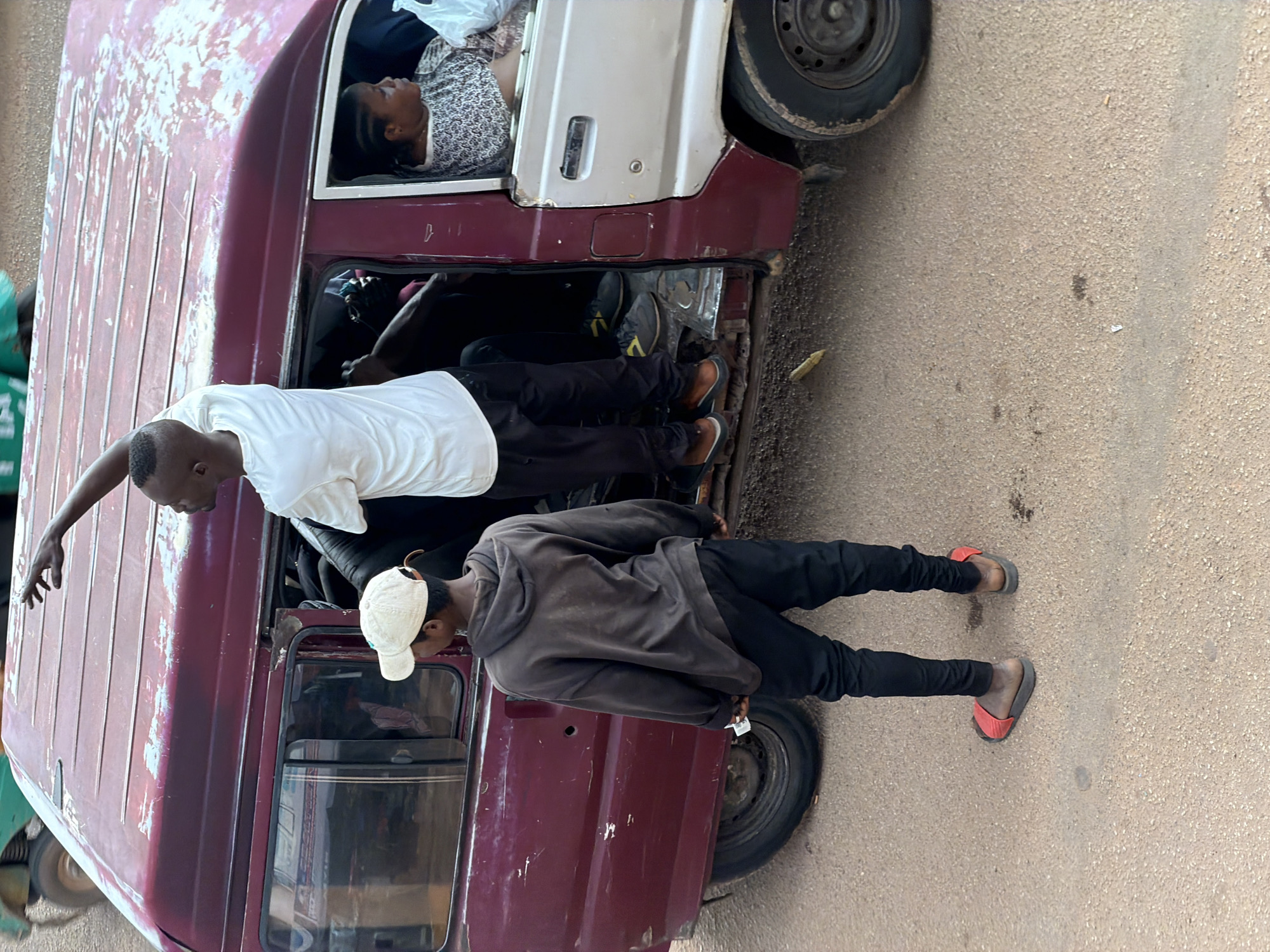 In Bukuru, Nigeria, two young men are visible on the exterior of a maroon van. One man, wearing a white t-shirt and black pants, is standing on the roof of the van, with his right arm extended upwards as if signaling or pointing. He is positioned on the right side of the van's roof. The second man, younger in appearance and wearing a beige baseball cap, a dark grey hooded sweatshirt, and black pants, stands to the left of the van, facing away from the camera and towards the vehicle's open doorway. He is wearing red slide sandals.
The van itself appears worn, with faded paint and visible rust spots. The side door on the left is ajar, revealing the interior of the van which is occupied by several people. A woman with braided hair, wearing a patterned top, is visible seated inside the van, looking outwards through the window on the right side of the vehicle. The van's wheels are visible, and it rests on a dirt or gravel road, indicative of an outdoor setting in Bukuru. The overall scene suggests a public transportation scenario, possibly a mini-bus, in motion or preparing for departure.