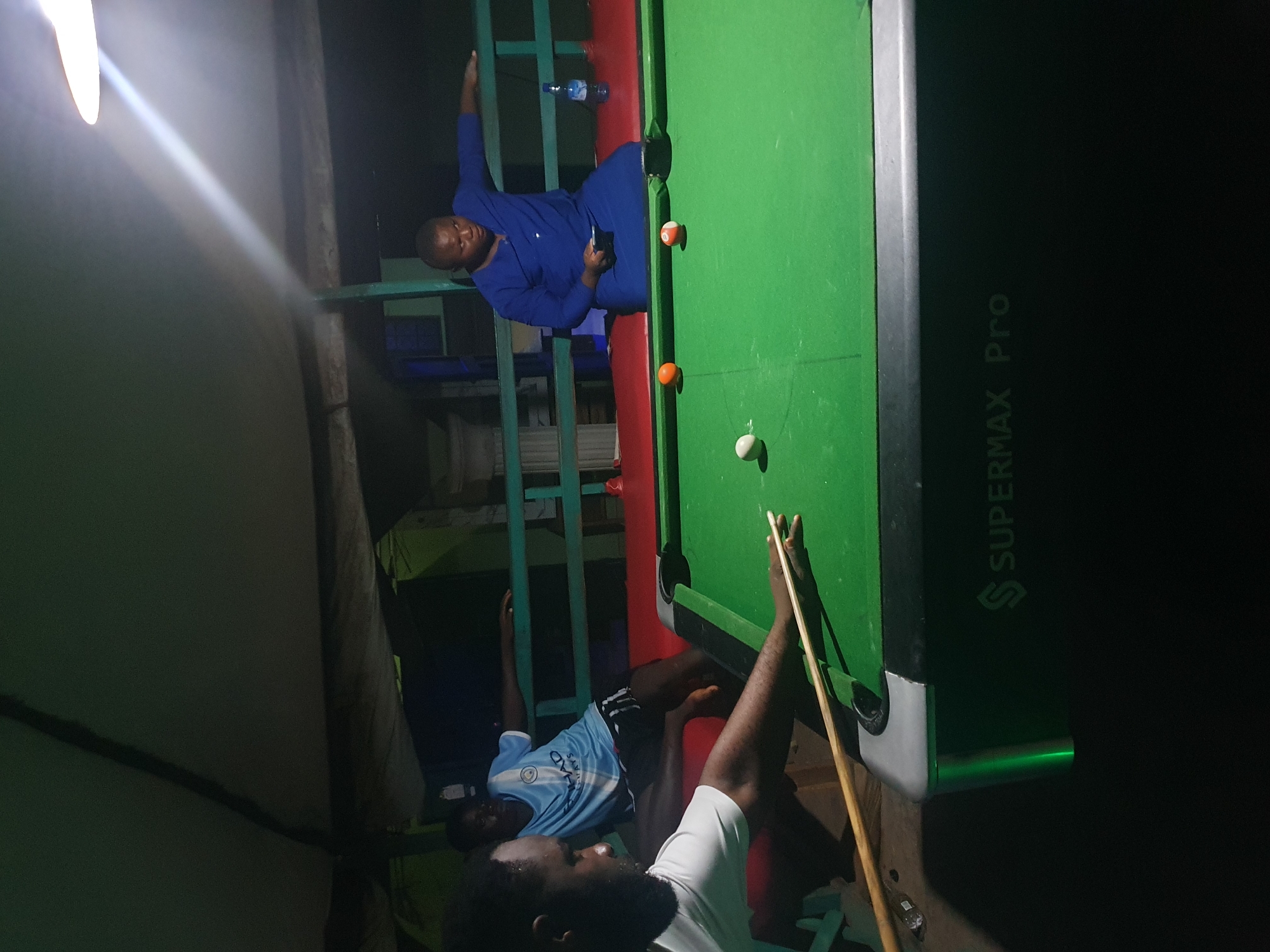 The image depicts an indoor pool hall in Lokoja, Nigeria, with three individuals present. A person in a white t-shirt is actively playing pool, positioned to the left of the frame and holding a cue stick. To their right, another individual, wearing a light blue Manchester City jersey and black shorts, is seated. In the background, a third person in a blue robe is seated and holding a cell phone, with a bottle of water on the railing behind them. The pool table is green with visible balls, including an orange ball, a white ball, and a ball with red and white markings. The environment appears to be a covered outdoor space or a dimly lit indoor area, indicated by the presence of a ceiling or canopy and a bright overhead light source. The floor is dark, and the background shows a railing and some structural elements. The pool table itself is branded "SUPERMAX Pro."