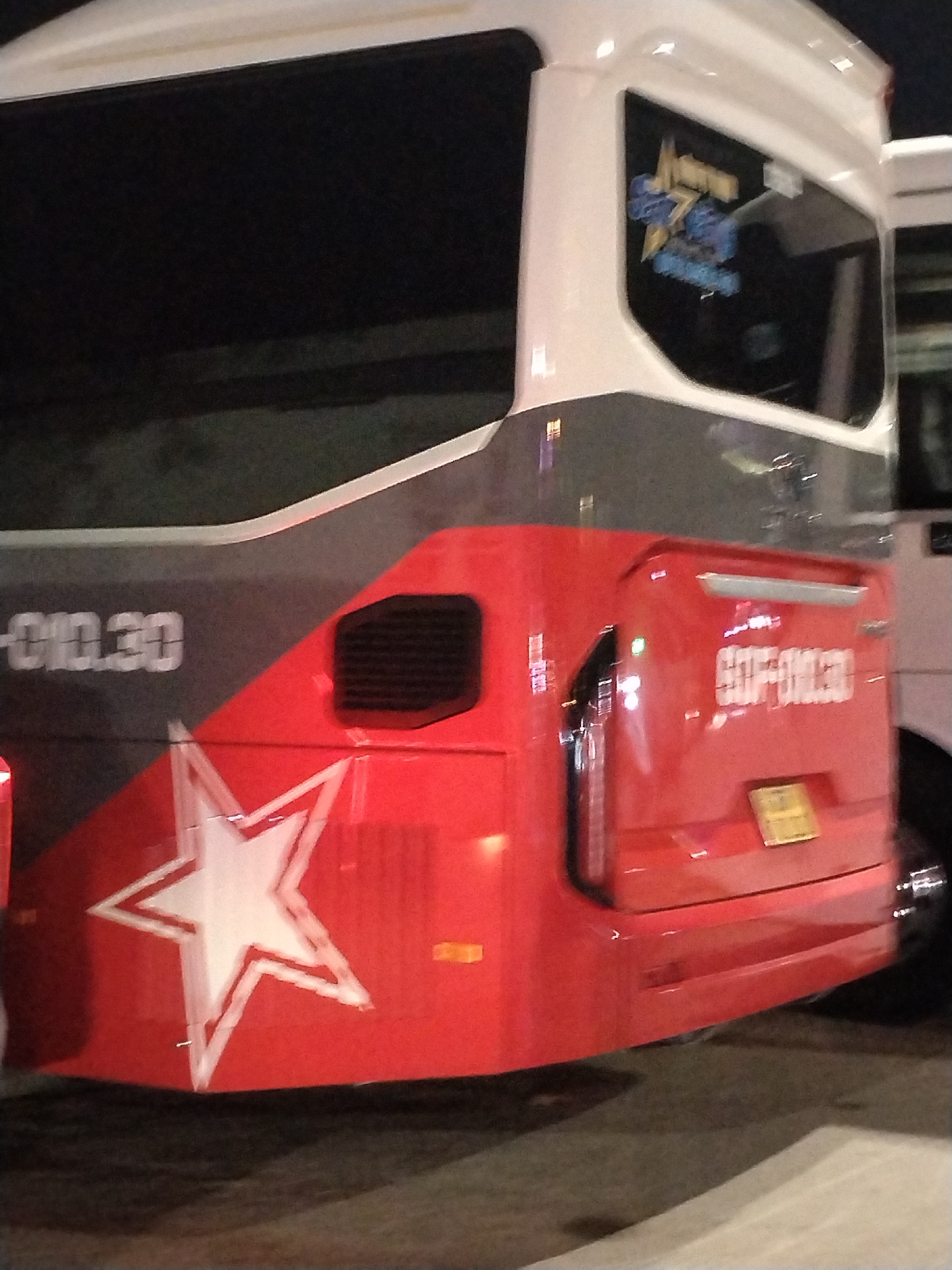 The image shows a close-up of the side of a red and gray bus. A white star outlined in red is visible on the red lower section of the bus. To the left of the star, the text "-010.30" is visible in white on the gray section. To the right of the star, on the red section, the text "90F-010.30" is visible in white. A black rectangular vent is located above and to the right of the star. On the upper white section of the bus, a blue and yellow sticker with a star design is visible. The scene appears to be outdoors at night, with some reflections from lights visible on the bus's surface. The image does not provide enough detail to determine the specific location within Bac Nha Trang Ward, Vietnam.