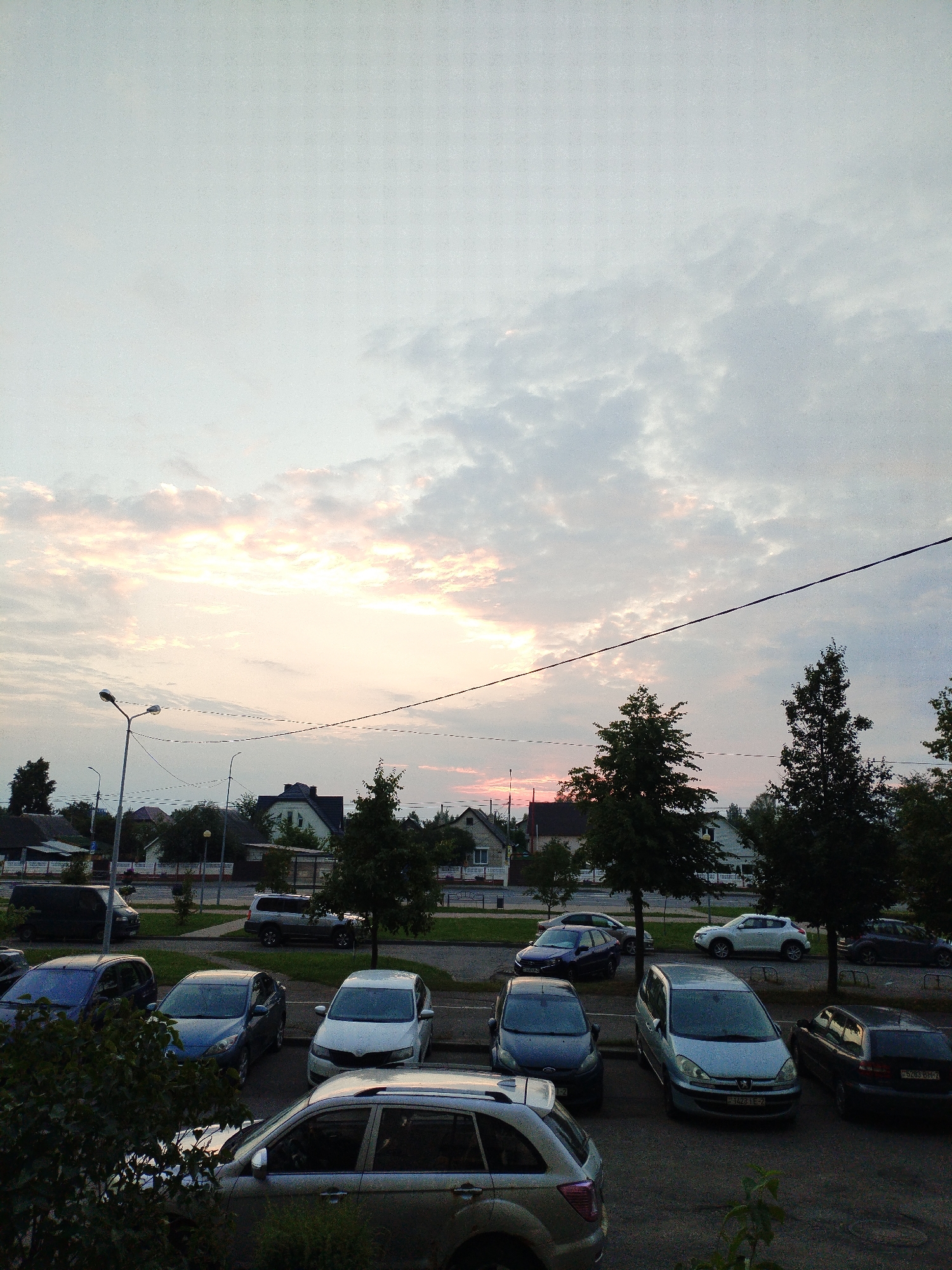 The image captures a view of a street in Polatsk, Belarus, at dusk. The sky is filled with clouds, with hints of pink and orange from the setting sun. A power line stretches across the frame. In the foreground, several cars are parked on asphalt. These include a silver SUV, a blue sedan, a dark hatchback, and a lighter colored sedan. Behind the parked cars, there are trees and houses with pitched roofs. Streetlights are visible on the left side of the image. The overall scene depicts a quiet residential or street area during the transition from day to night.
