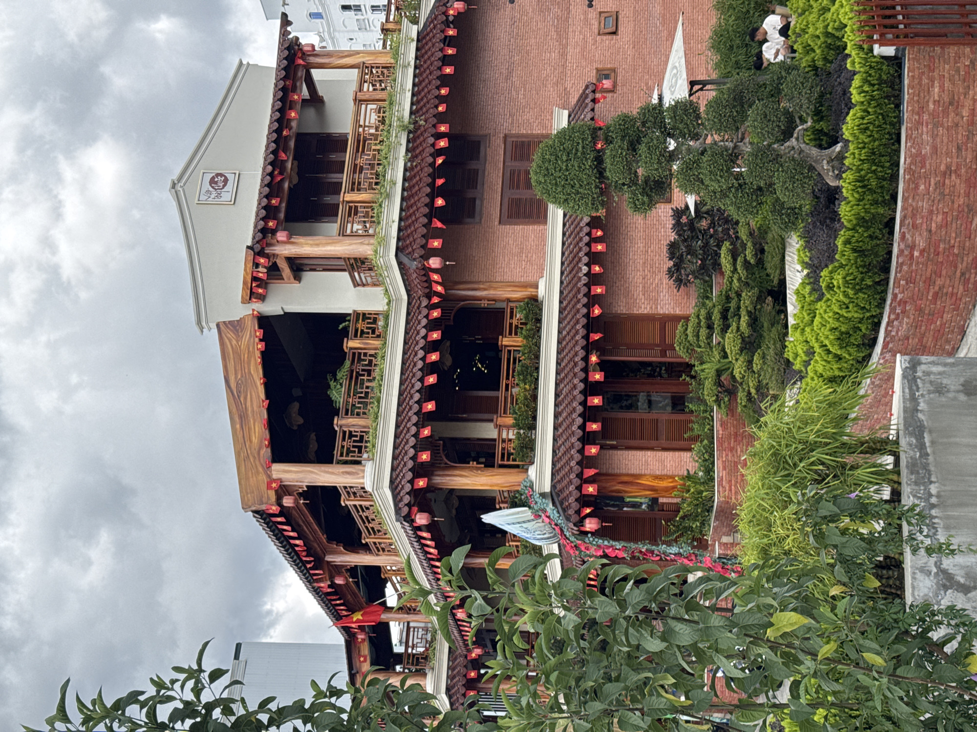 The image captures a multi-story building in Da Lat, Vietnam, characterized by its traditional Vietnamese architecture. The building has wooden pillars and beams, dark brown tile roofs, and is adorned with small Vietnamese flags. A white-painted section with a sign in Asian characters is on the uppermost part of the structure. The building's facade is primarily brick, with wooden shutters on the windows. Lush greenery surrounds the building, including bonsai-like trees and shrubs. A cloudy sky is visible in the background, with some glimpses of adjacent urban structures.