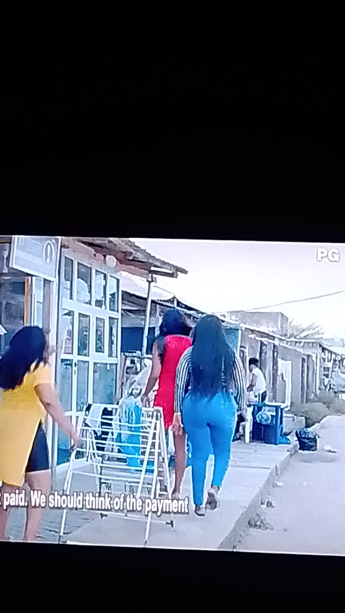 The image depicts a street scene in Lokoja, Nigeria. In the foreground, a portion of a white clothes drying rack is visible. Three women are walking away from the camera down a paved sidewalk. The woman on the left is wearing a yellow top and dark shorts. The middle woman is wearing a red dress. The woman on the right is wearing a striped top and bright blue leggings. Behind them, buildings with corrugated metal roofs and glass-fronted facades line the street. The environment appears to be a commercial area with small shops. Text overlays indicate "PG" in the upper right and "t paid. We should think of the payment" at the bottom left.