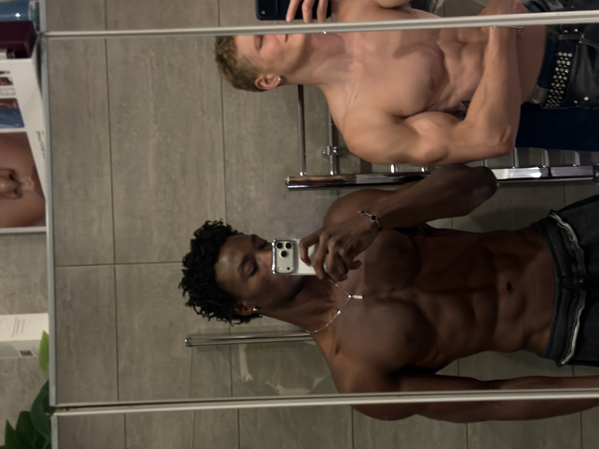The image presents two shirtless young adult males reflected in a large mirror, situated within an indoor environment featuring light grey tiled walls and a visible metal towel warmer. A shelf above the mirror displays a white bottle and indistinct printed material. The male on the left, exhibiting a dark skin tone and short, dark, curly hair, possesses a muscular physique with prominent abdominal musculature. He is dressed in dark athletic shorts and accessorized with a silver chain necklace featuring a white pendant, a small silver stud earring in his left ear, and a bracelet on his left wrist. He holds a silver-colored smartphone, characterized by a prominent camera module, in his right hand, positioning it to capture a mirror selfie while obscuring his lower face. The male on the right, with a lighter skin tone and short, curly blond hair, also demonstrates a muscular build with defined biceps and abdominal muscles. He wears black trousers secured by a black studded belt and a small silver hoop earring in his left ear. His face is partially obstructed by the mirror's edge and a dark blue smartphone held in his right hand. Both individuals are engaged in posing for the photograph. The image does not contain identifiable markers to confirm its location in London, United Kingdom.