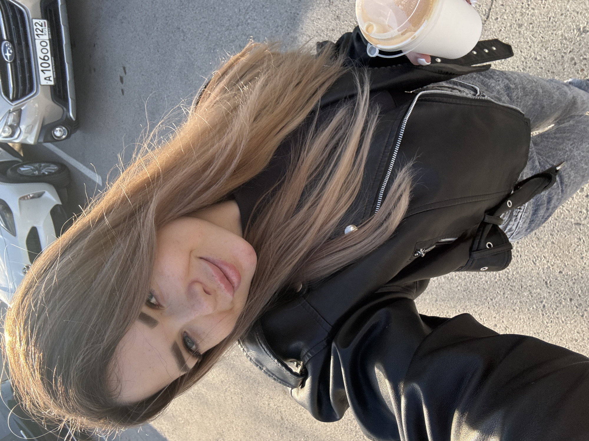 A woman takes a selfie while standing outside, holding a coffee in her right hand. She is wearing a black leather jacket and gray jeans. Parked vehicles are visible on the left. The location is the city of Unknown, Unknown.