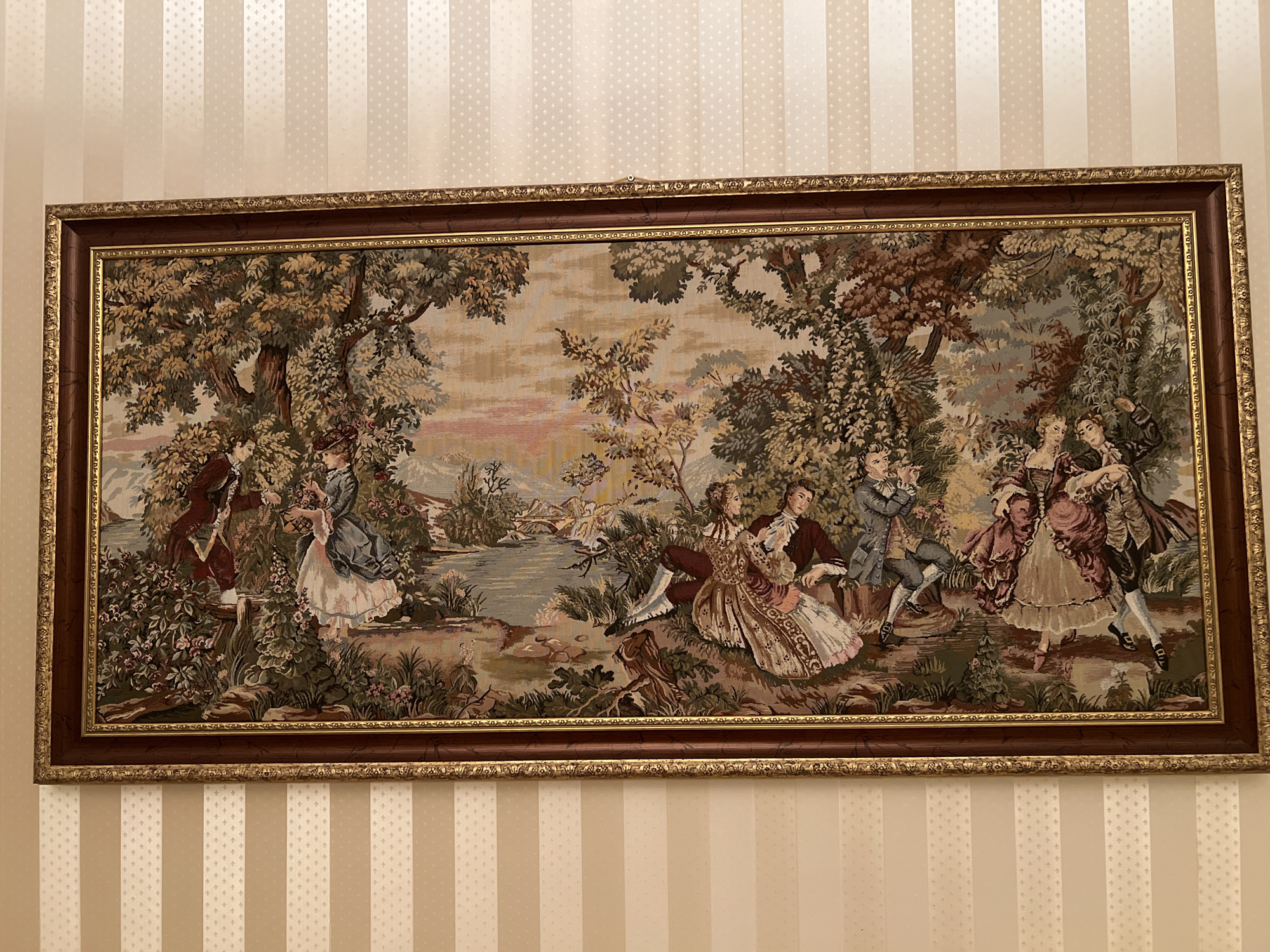A horizontal, framed tapestry hangs on a wall with cream and tan vertical stripes. The tapestry depicts a pastoral scene with several figures in 18th-century attire. In the foreground, a man and woman sit together, with the woman wearing an ornate pink and cream dress and the man in a dark jacket and breeches. To their left, a woman in a blue dress stands near a body of water. Further left, another woman in a red dress and another man in a dark jacket are partially visible. On the right side of the tapestry, several figures are dancing, including a man playing a flute. The background features trees, a river, a bridge, and distant hills under a pale, cloudy sky. The tapestry is framed by a dark wooden inner frame and a more ornate gold outer frame. The location is the Bender Fortress Museum in Bender, Moldova.