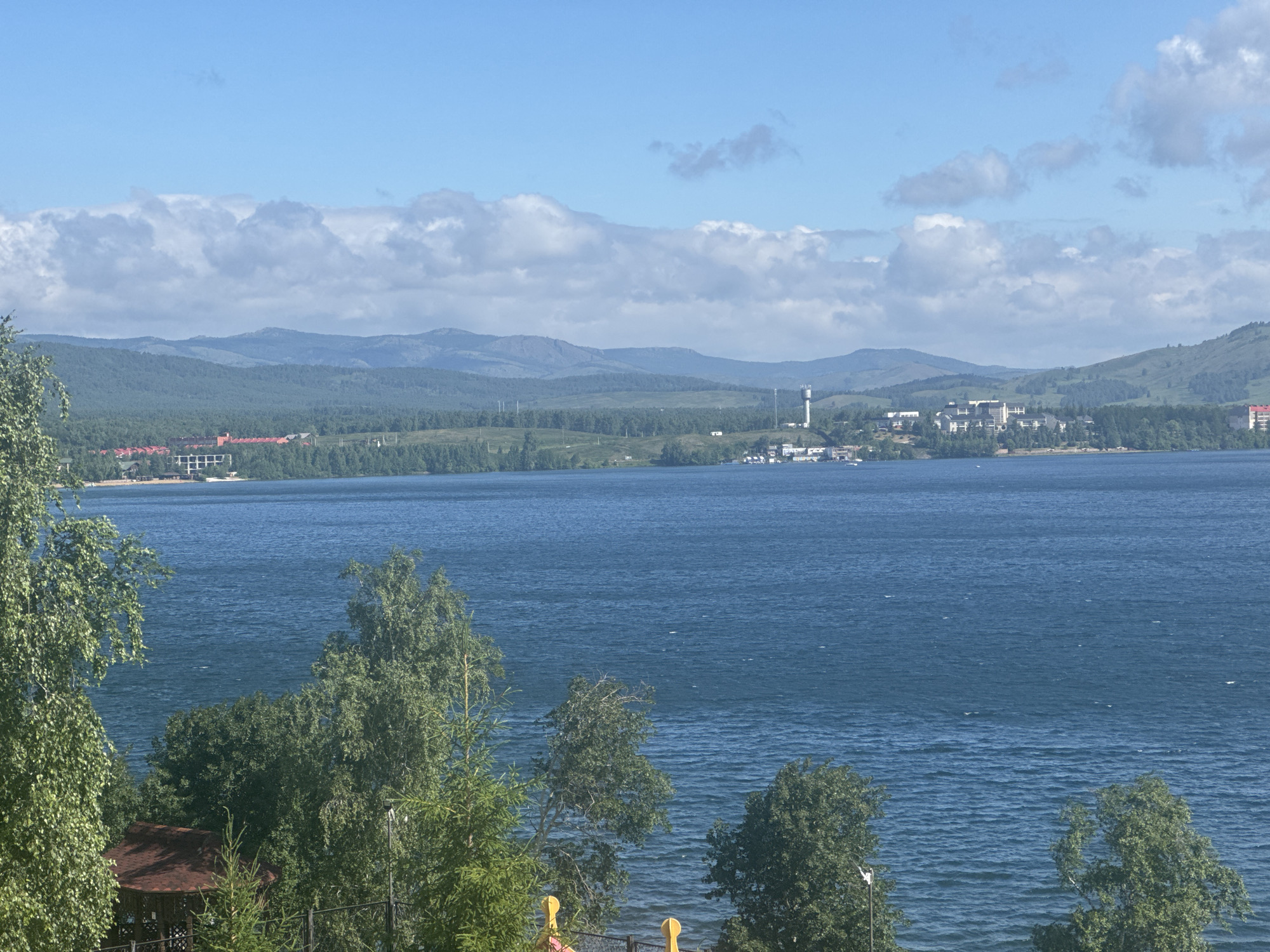 The image captures a wide-angle view of a serene lake, likely Lake Yakty-Kul in the Republic of Bashkortostan, Russia, on a clear, sunny day. The foreground is dominated by lush green trees and foliage, with a glimpse of a small, reddish-brown gazebo with a conical roof. Beyond the trees, the expansive, deep blue lake stretches towards the horizon. Across the lake, a line of buildings is visible, including what appears to be a hotel or resort complex with red roofs and a cluster of white buildings. In the distance, rolling, tree-covered hills and mountains form a picturesque backdrop, with layers of blue and green receding into the hazy distance. The sky is a bright, clear blue, punctuated by scattered white clouds, some of which form a soft band across the sky above the mountains. The overall impression is one of natural beauty and tranquility, with signs of human habitation integrated into the landscape.