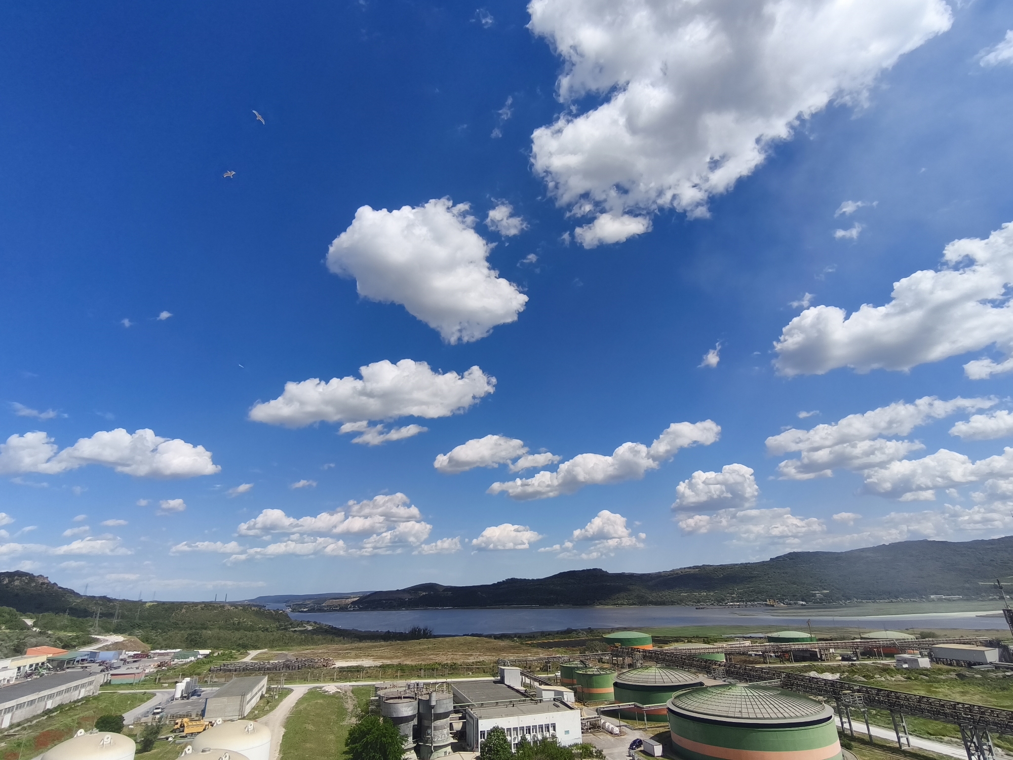 An elevated perspective captures a sunny day with a blue sky dotted by white cumulus clouds. In the distance, a body of water is framed by undulating hills. In the foreground, industrial architecture, including storage tanks with green and orange stripes, is juxtaposed with green fields and an assembly of buildings with flat roofs. Two birds are seen in the sky. The location is the city of Unknown, Unknown.