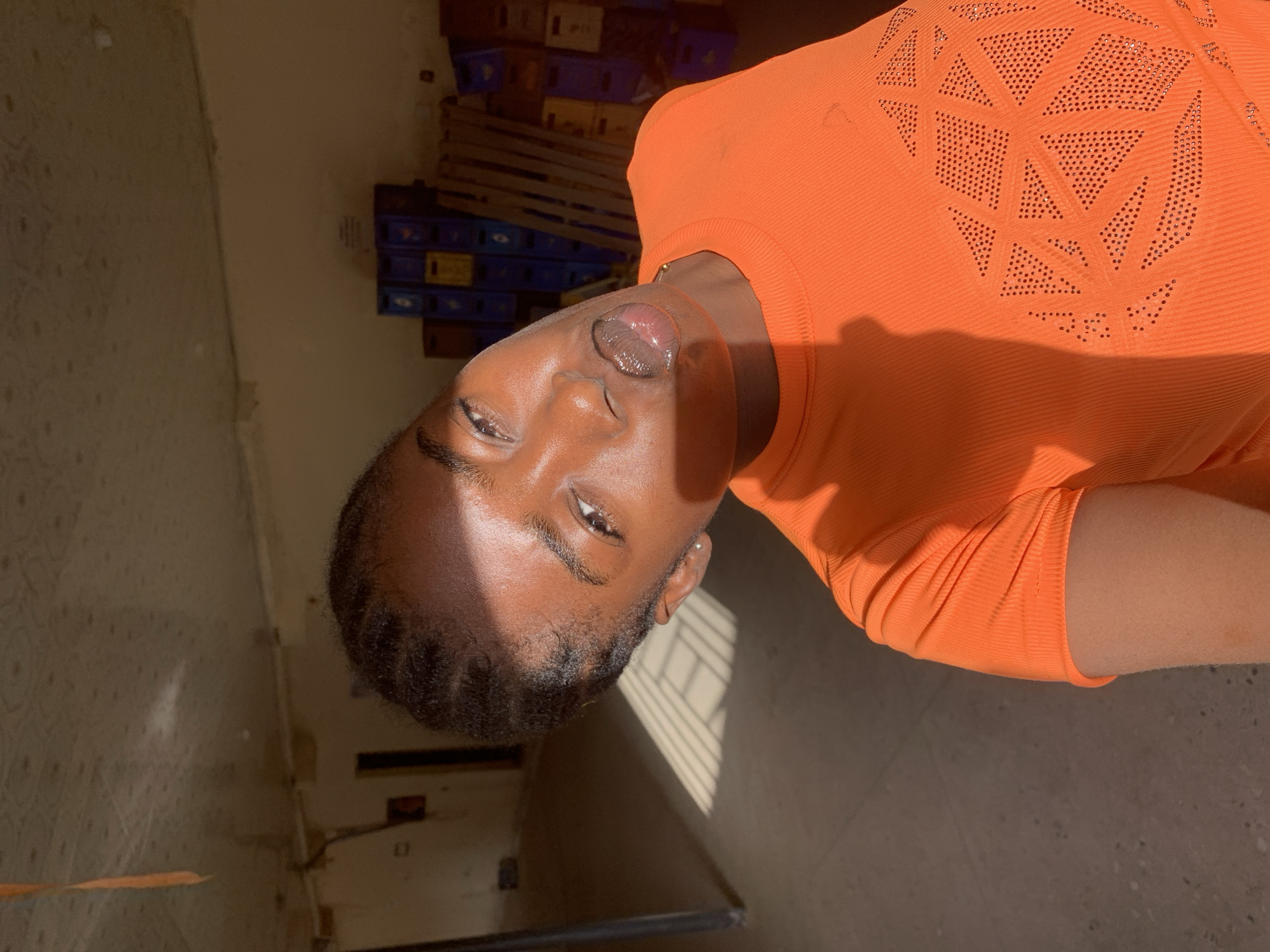 A young dark-skinned person with short, dark, braided hair and an orange top is indoors. The person is angled to the left. The background includes a wall, a floor, window shades, and stacks of blue and yellow plastic crates. The location is in New Stadium, Nigeria.