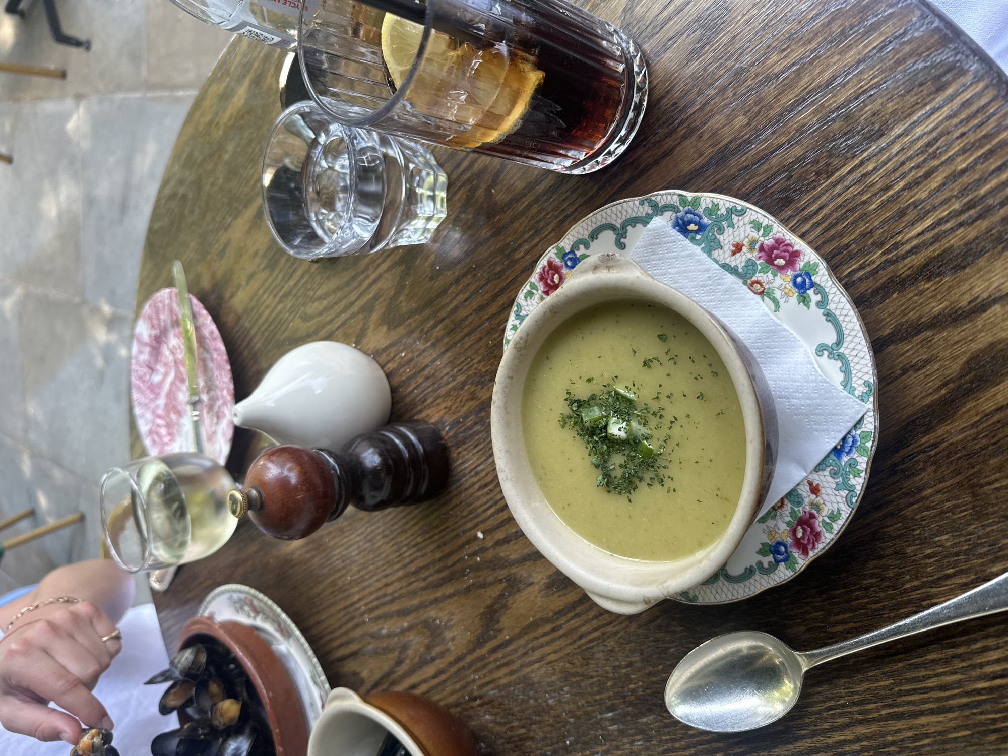 A top-down view captures a meal setting on a wooden table. A bowl of green soup, garnished with herbs, sits on a floral patterned plate with a white napkin. A silver spoon is placed beside the plate. Next to these are a small plate, a white sauce container, a wooden pepper grinder, a glass of white wine, and a dish of mussels. A tall glass of dark liquid with a lemon slice and a glass of water are also visible. A hand is seen picking up a mussel. The location is Unknown, Unknown.