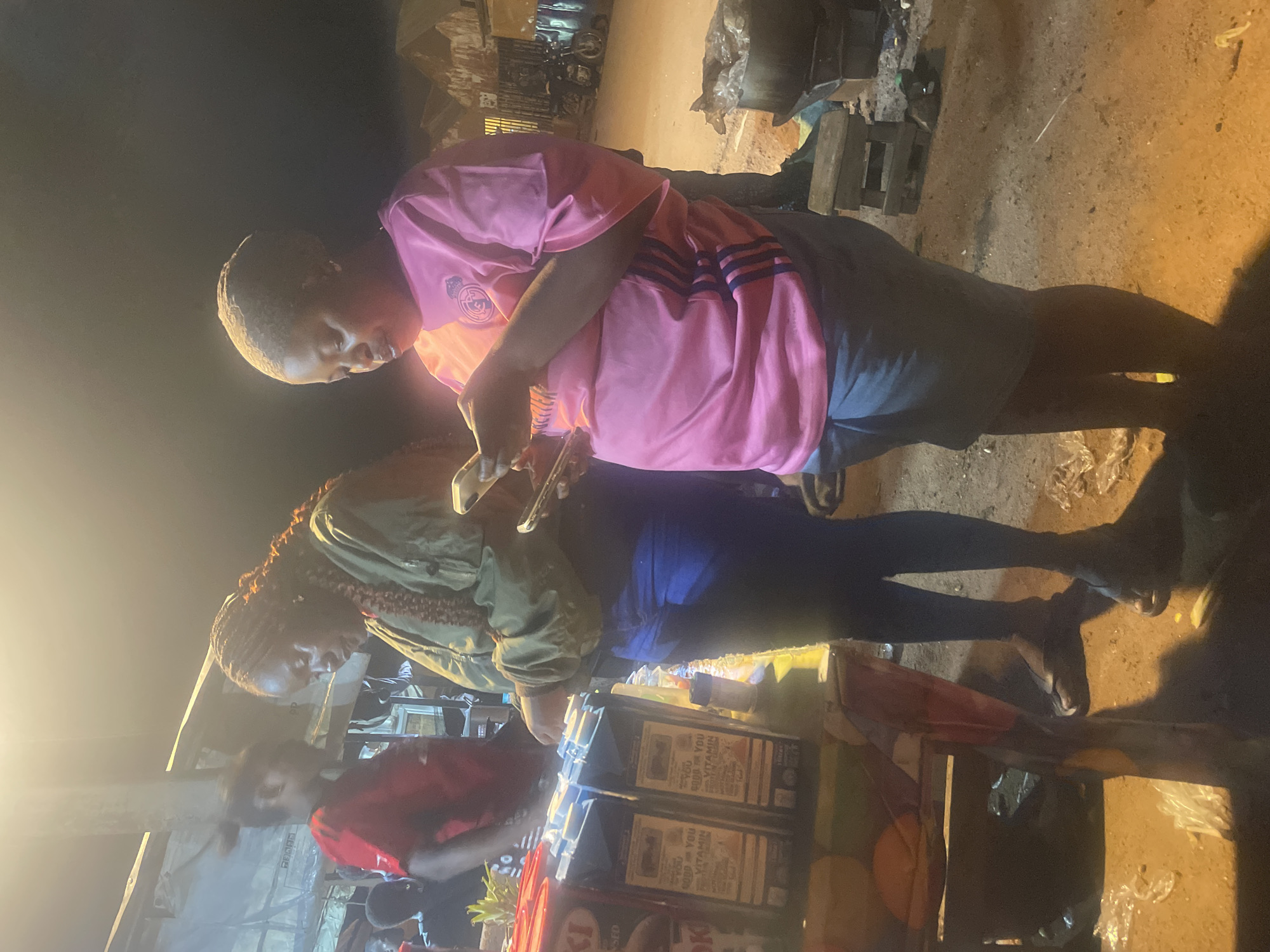 Two women are standing at a market stall in Du, Nigeria. The woman on the left is wearing a green jacket and blue jeans, and she is looking at her phone. The woman on the right is wearing a pink t-shirt and grey shorts, and she is also looking at her phone. The market stall is filled with boxes of juice. There are other people in the background, but they are out of focus. The scene is dimly lit, suggesting it is nighttime.