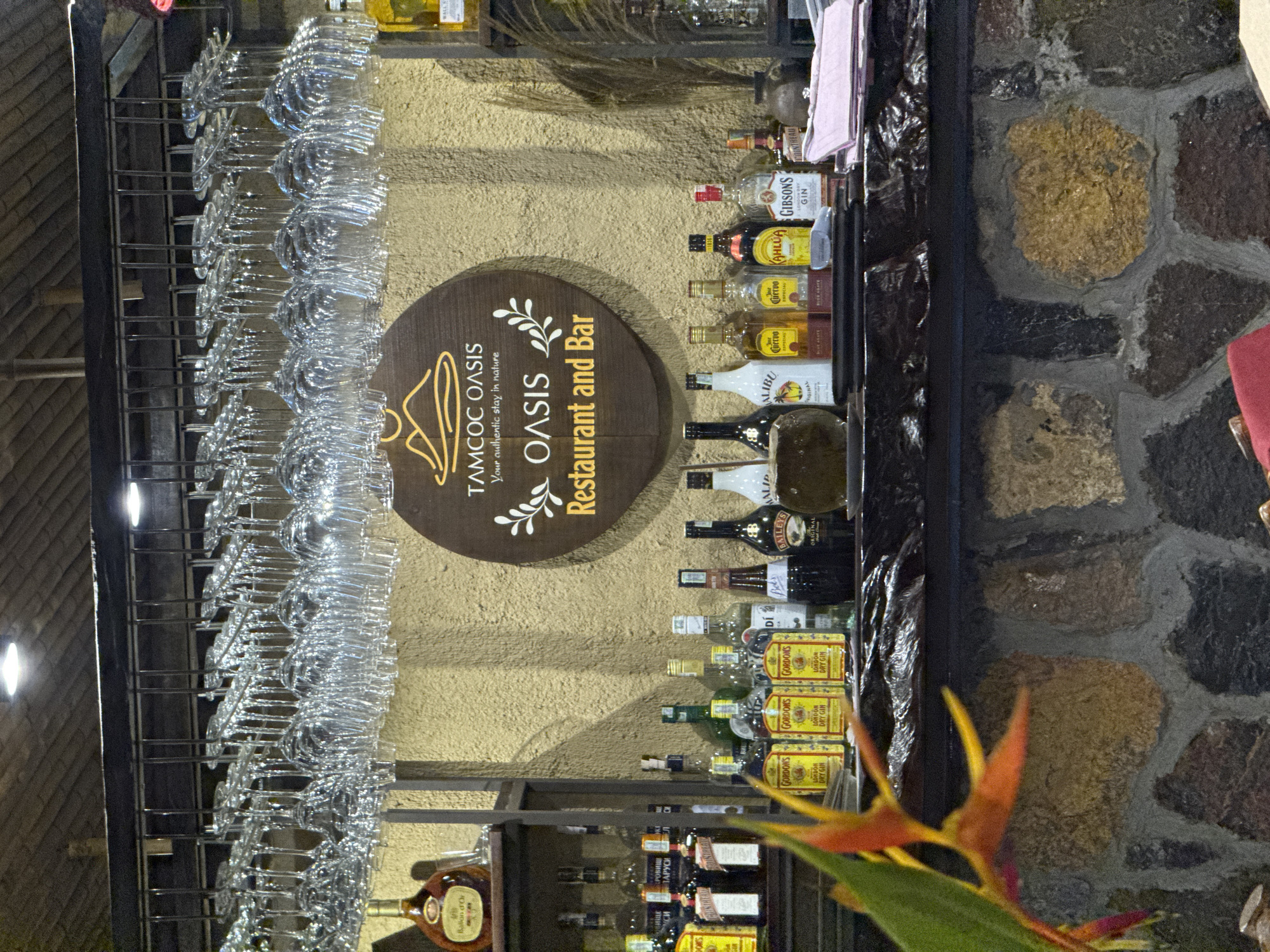 The image depicts a bar counter in the city of Nam Hoa Lư Ward, Vietnam. Suspended above the counter are numerous wine glasses. The bar itself is constructed from stone and is stocked with various bottles of alcohol. Notable brands visible include Gordon's London Dry Gin, Malibu, Jose Cuervo, Bailys, and Cointreau. A decorative sign for "TAMCOC OASIS Restaurant and Bar" is prominently displayed on the wall behind the counter. In the foreground, a tropical flower with orange and yellow petals adds a splash of color to the scene. The lighting suggests an indoor setting, likely in the evening.