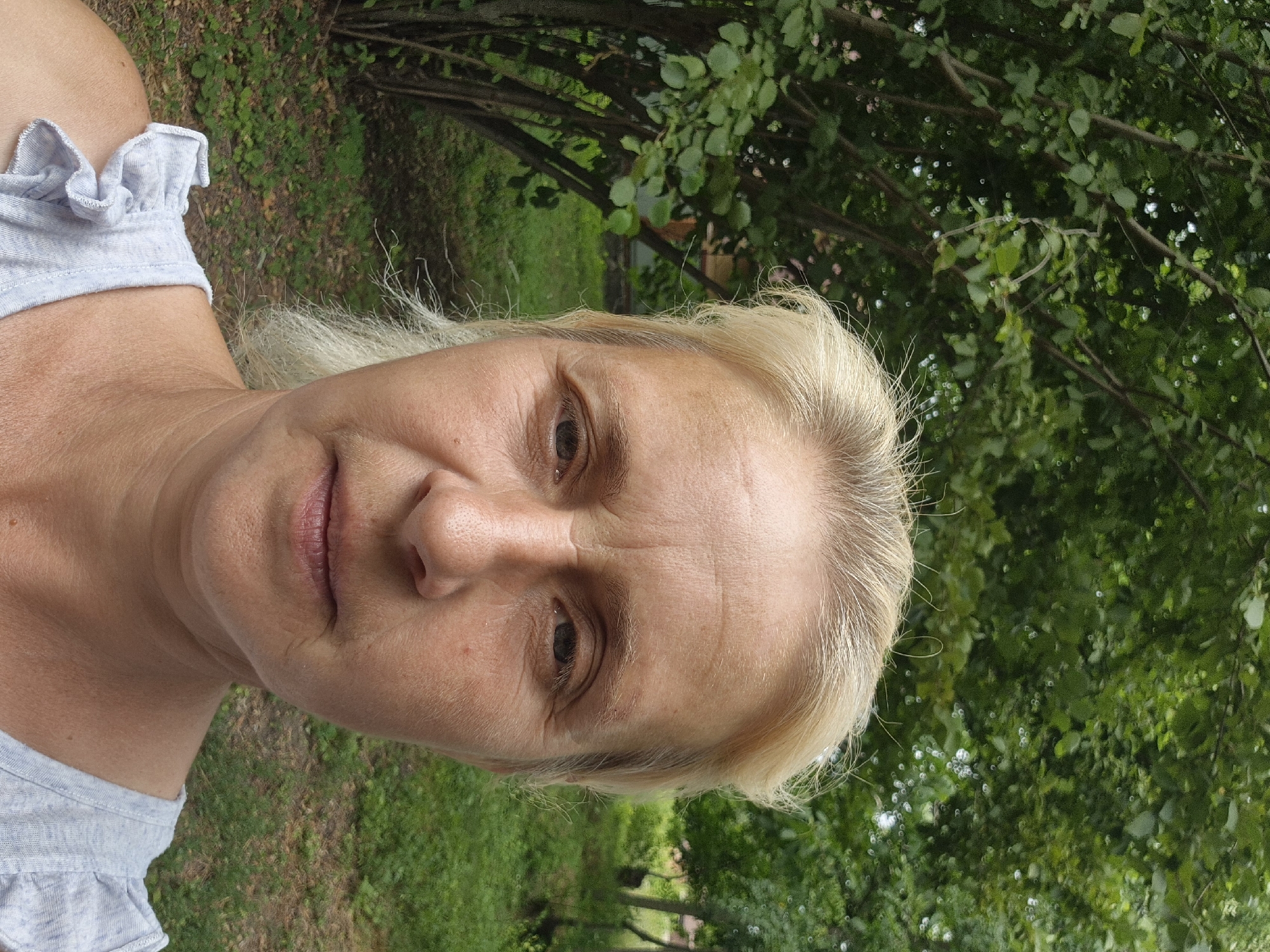 A medium shot shows a woman with blonde hair and fair skin, looking directly at the camera. She has wrinkles on her forehead and around her eyes. The woman is wearing a light gray ruffled shirt. She is outdoors, with lush green trees and foliage in the background. The environment suggests a natural setting, possibly a park or a garden. The image does not contain any specific identifiable landmarks or features that would pinpoint its location within Sozonivka, Ukraine.