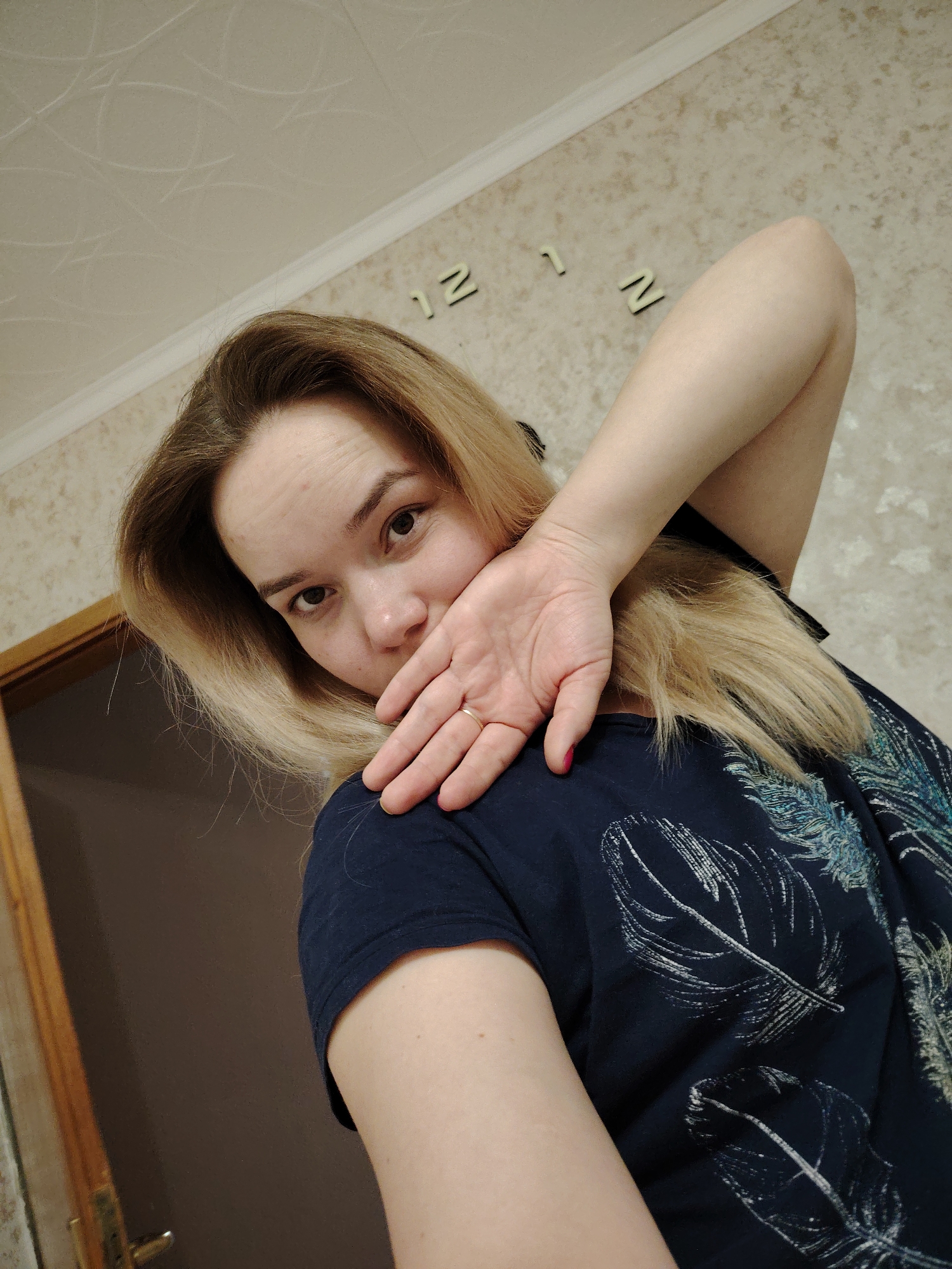 A woman with blonde hair and fair skin is looking at the camera with a slightly playful expression. She is wearing a dark blue t-shirt with a light-colored feather pattern. Her right hand is raised to her face, with her fingers partially obscuring her mouth. She is wearing a ring on her ring finger and nail polish on the little finger of the hand that is obscuring her face. The background shows a wall with a beige wallpaper that has a subtle geometric pattern, as well as letters "12" on the wall. A door with a wooden frame is partially visible on the left side of the frame. The image appears to be taken in an indoor setting, possibly a home. Although the location Марковский, Russia cannot be verified from the information in the image, if this is accurate, it would be a residential setting in that location.