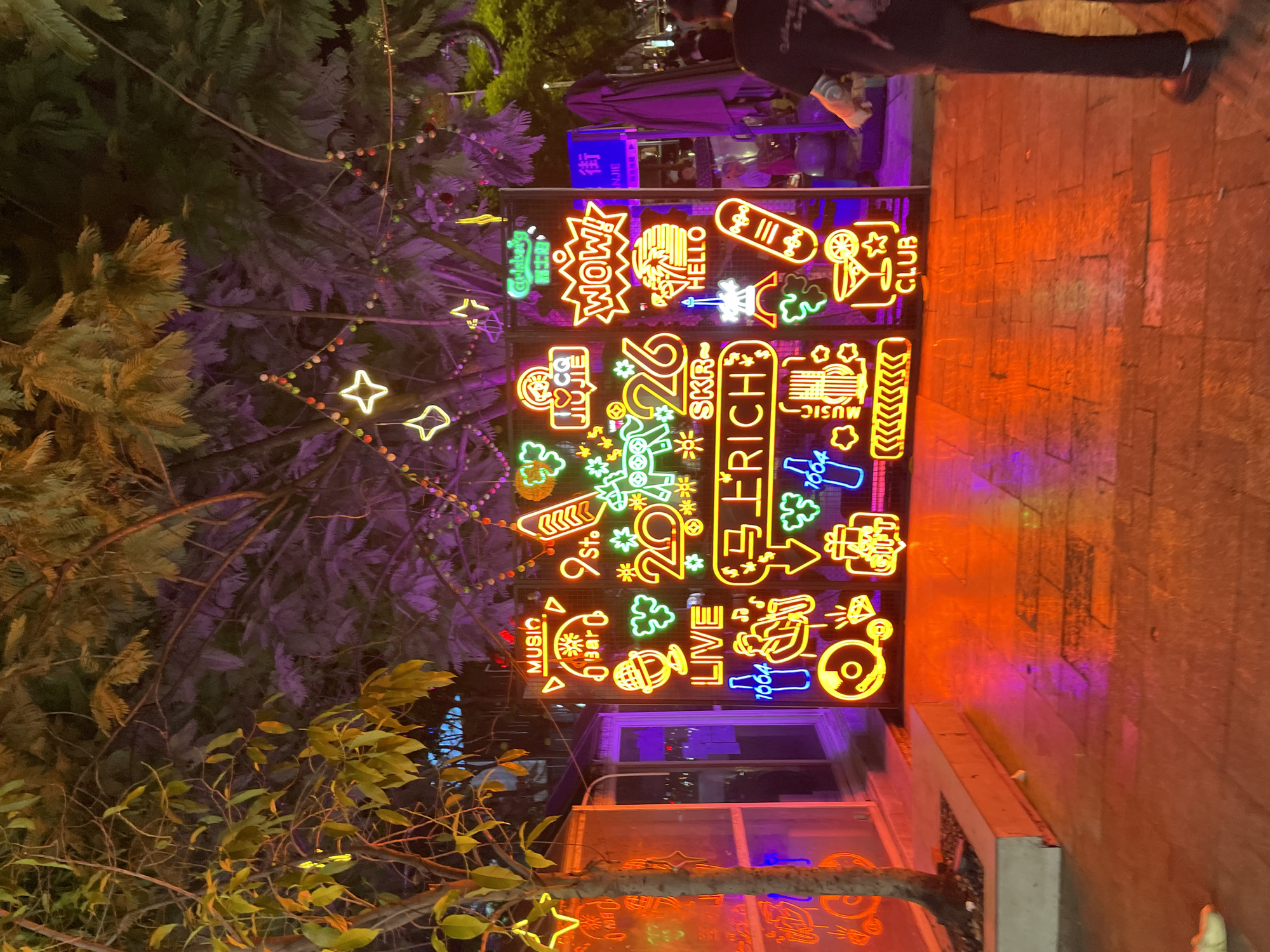 This is a nighttime, medium shot of a vibrant street scene in Liangjiang, China, illuminated by numerous neon signs. The signs are arranged on a metal frame and display a variety of graphics and text, including musical instruments, drinks, people, and words like "MUSIC," "LIVE," "WOW!," "HELLO," and "CLUB." The ground is wet, reflecting the colorful lights. In the background, trees are visible, some adorned with small decorative lights. A person is partially visible on the right side of the frame, facing away from the camera. To the left, a tree with green leaves stands next to a translucent structure with orange and purple lighting. The overall atmosphere is lively and festive, characteristic of a nightlife district.