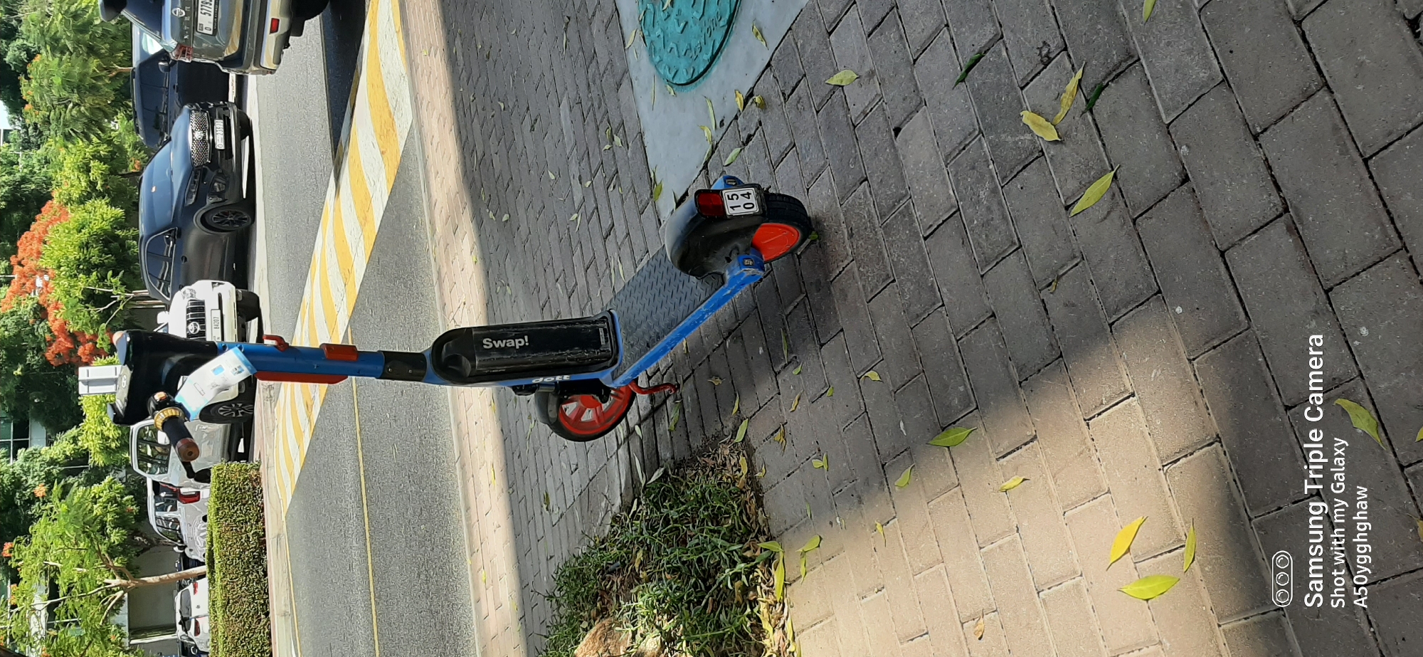 An electric scooter is parked on a brick sidewalk next to a street in Dubai, United Arab Emirates. The scooter is blue and black with red accents on the wheels and a license plate that reads "15 04". It has a black platform and handlebars. In the background, several cars are parked along the street, including a white SUV and a black SUV. Trees with green and orange foliage are visible behind the cars. The ground is paved with interlocking bricks and scattered with fallen leaves. The lighting suggests it is daytime with shadows cast by the scooter and trees. A white and blue sticker is attached to the scooter's handlebar.