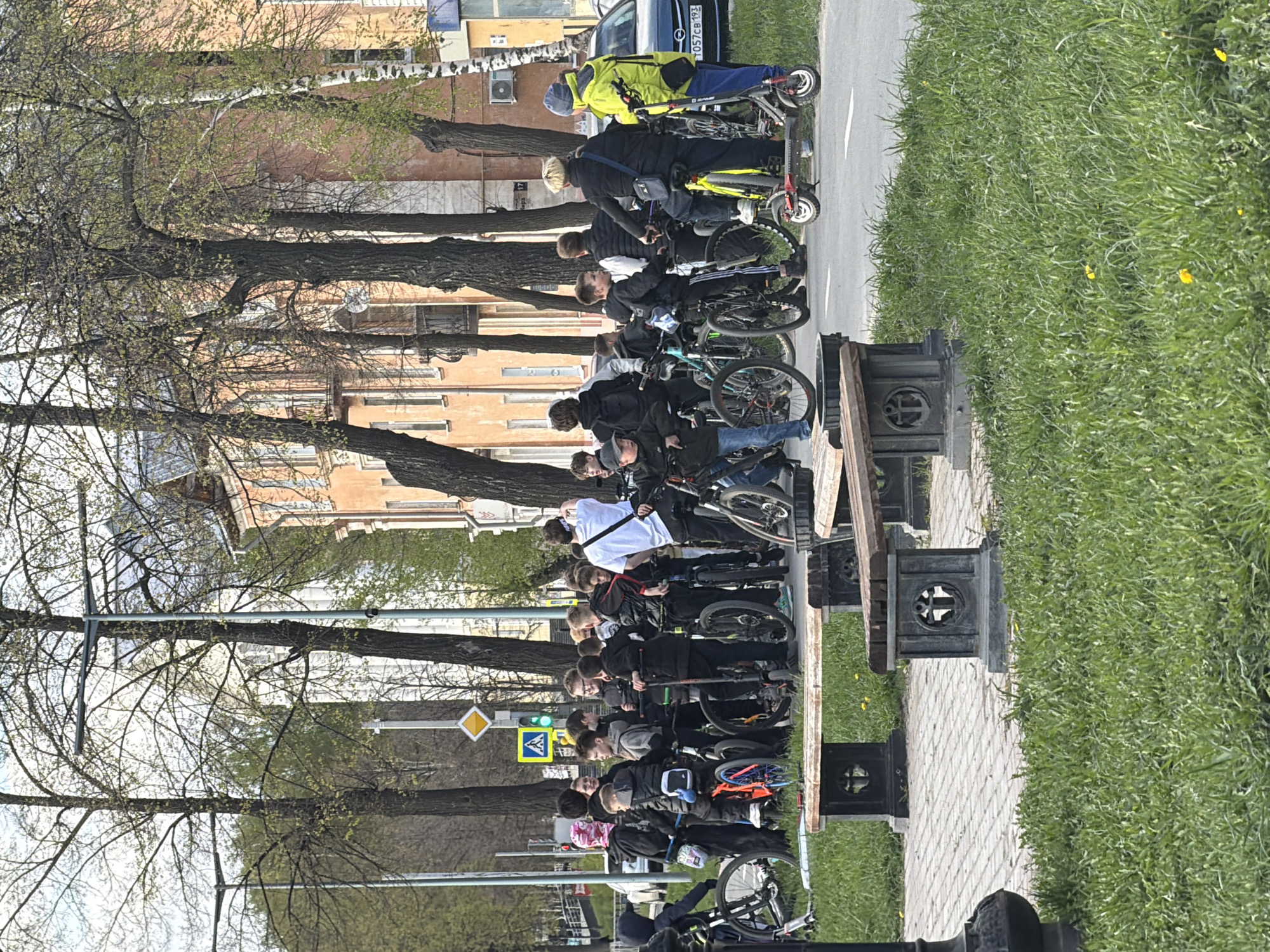 A group of approximately 15 people on bicycles and scooters are stopped on a sidewalk in Votkinsk, Russia, near a street with a car and a pedestrian crosswalk with a signal light. A line of benches are positioned along the sidewalk, adjacent to a patch of grass. Behind the sidewalk, there are buildings and trees.