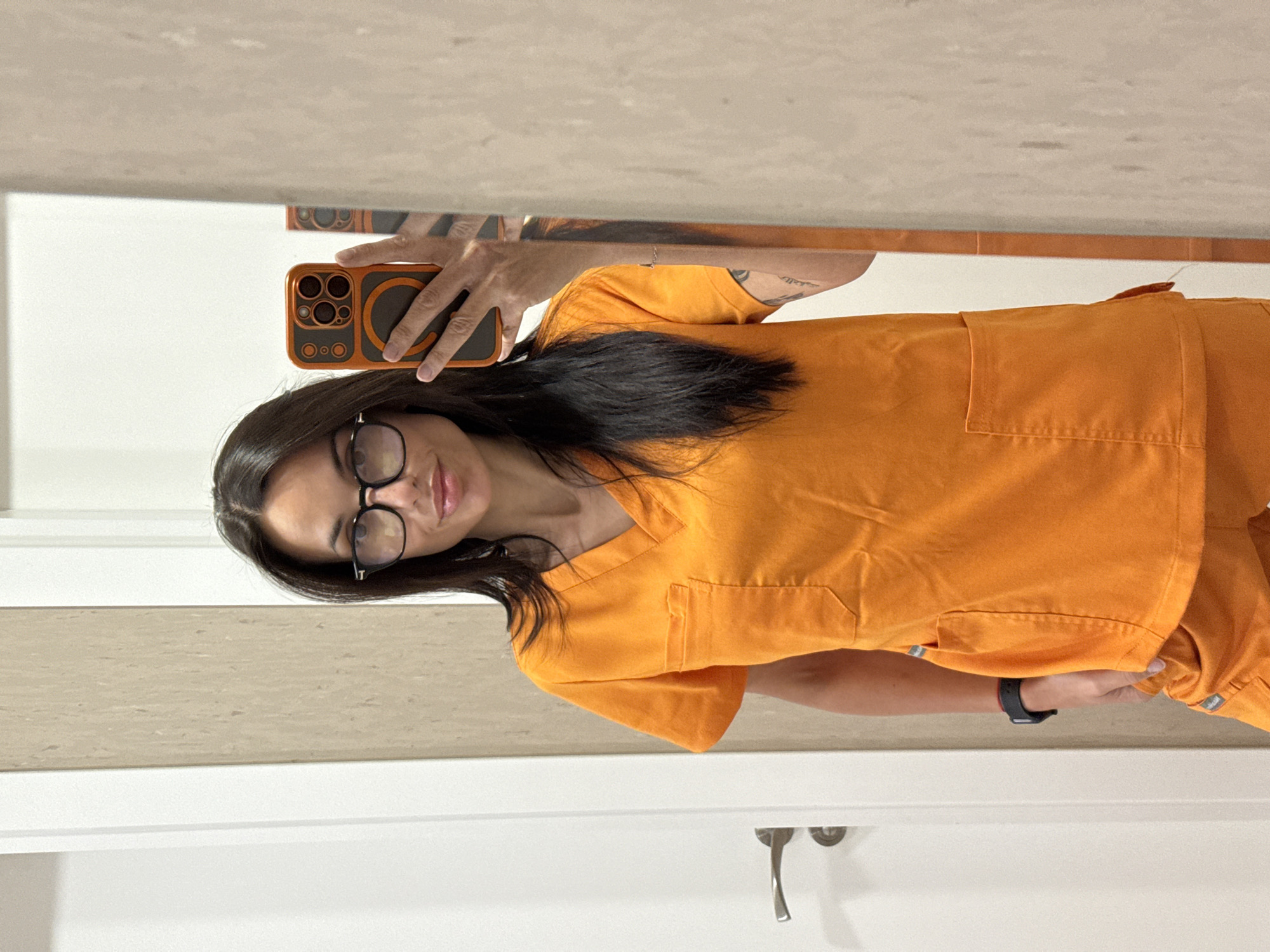A woman with long dark hair, wearing glasses and orange scrubs, is taking a selfie in a mirror. She is holding an orange iPhone with a black case in her right hand. A white wall and a light-colored counter are visible behind her and in the reflection. A silver faucet is visible near the bottom of the frame. The location is not discernible. No specific information related to Pleven, Bulgaria, is discernible in the provided image.