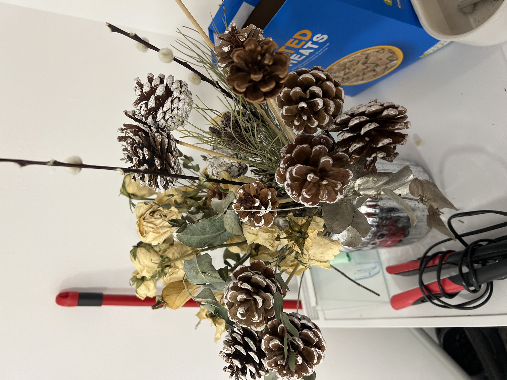 The image depicts a floral arrangement containing pine cones, dried roses, and other greenery, set against a white surface. A box of "Roasted Treats" and a pair of red-handled pliers are also visible. The exact location is Epsom and Ewell, United Kingdom. There are no people visible. The scene is a still-life arrangement of objects.