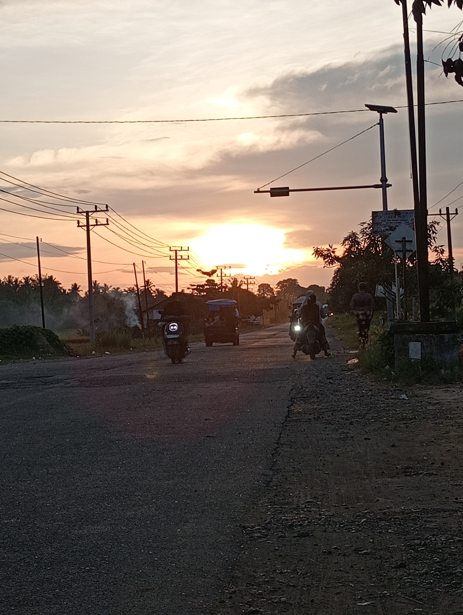 The image shows a street scene in Lhokseumawe, Indonesia, at dusk. The sky is bright from the setting sun. Overhead power lines and utility poles line both sides of the road. Several vehicles are present: a "becak," a small three-wheeled motorized vehicle, and two motorcycles traveling along the road. A person walks on the shoulder on the right. The road is paved, with a rough, gravelly shoulder on the right. Smoke can be seen on the left side of the road near some vegetation, possibly from burning. A street lamp with a solar panel is on the right.