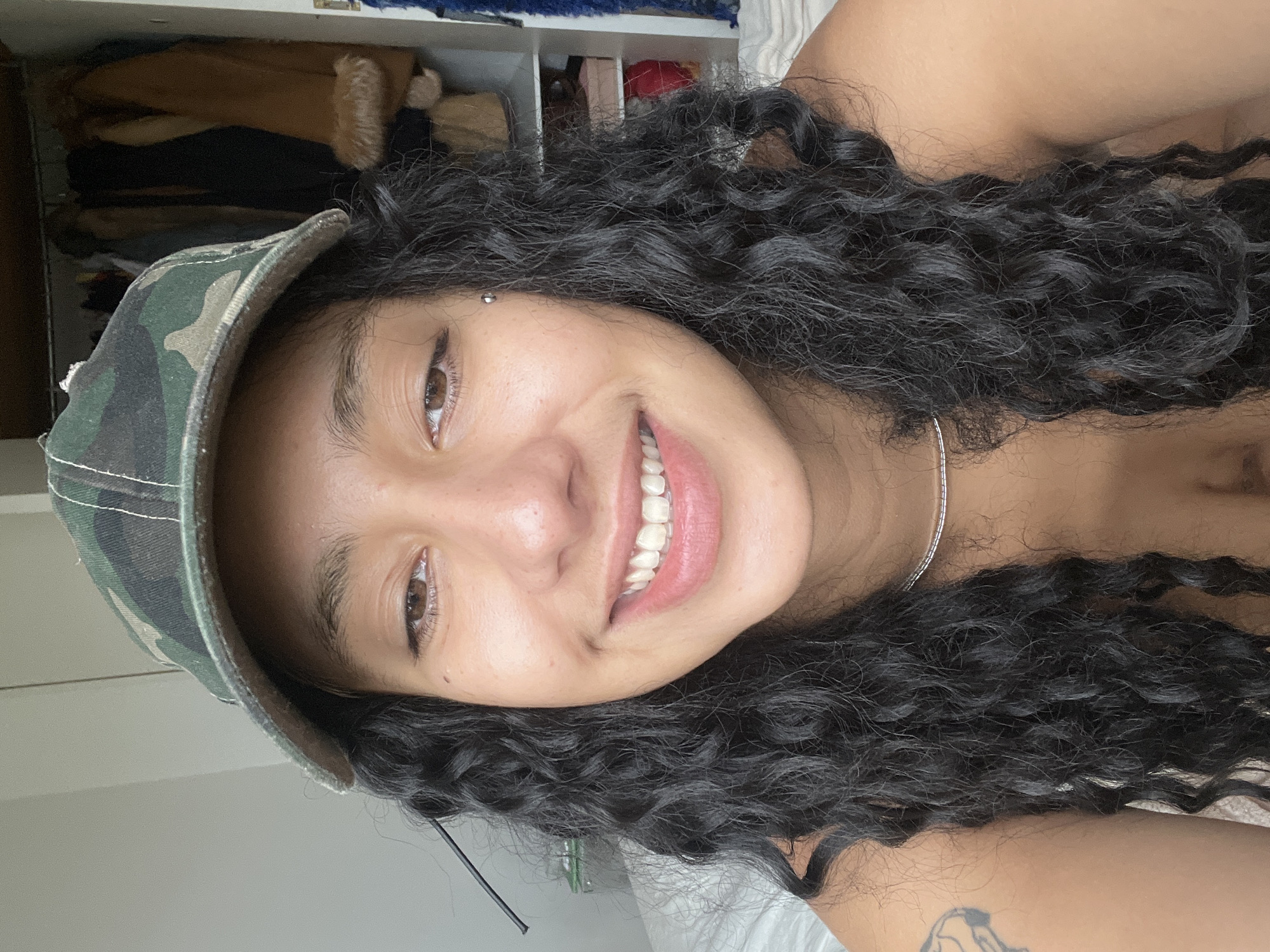 A close-up, rotated image depicts a woman with dark, curly hair and a camouflage baseball cap. She has brown eyes, light skin, and is smiling, revealing her teeth. She appears to have a piercing between her eyebrows. A thin silver necklace is visible. A partial tattoo is seen on her upper arm. In the background are blurred shelves containing various items of clothing and other objects. No details suggest the location is Buenos Aires, Argentina.