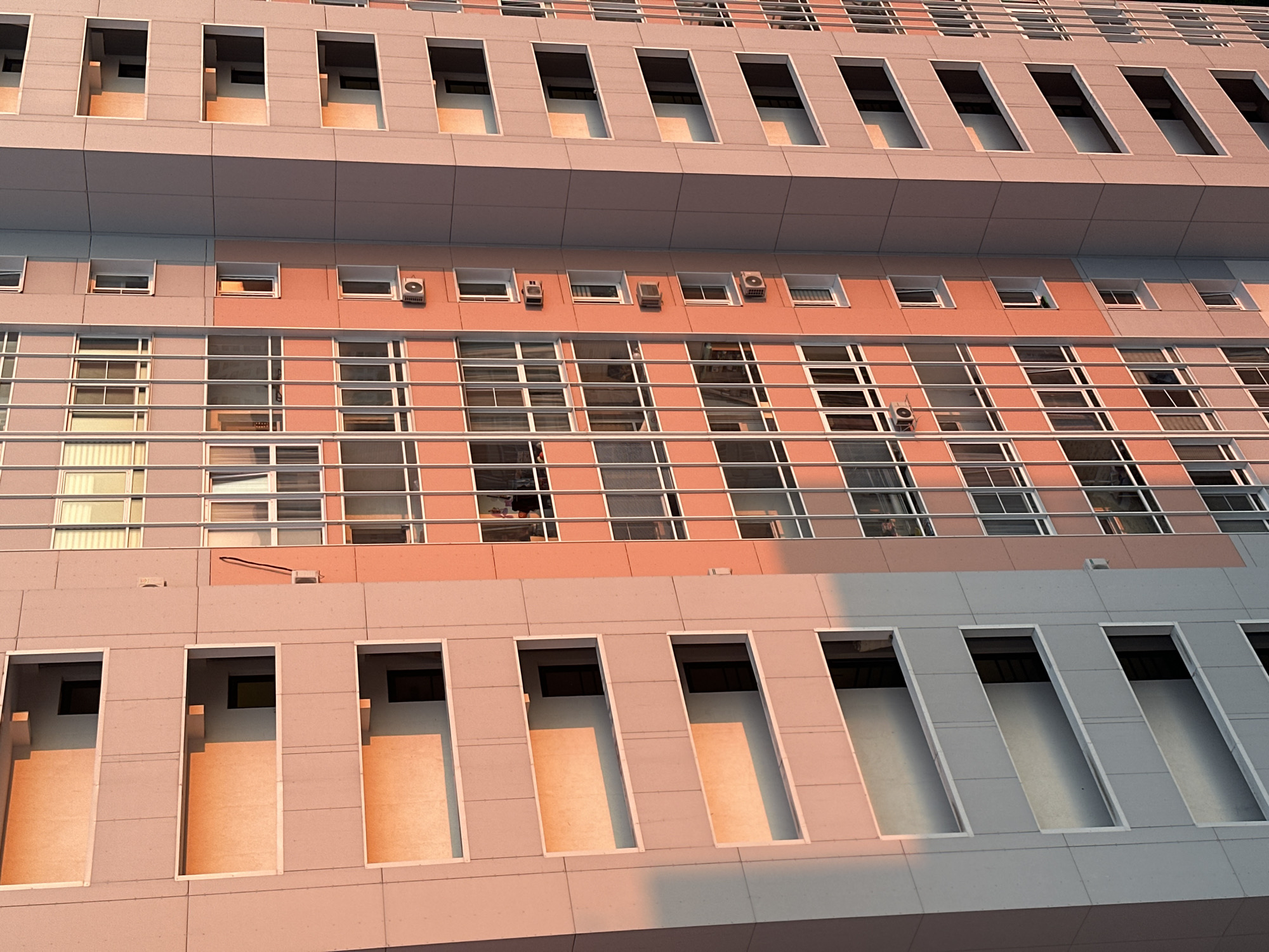 The image depicts a close-up view of a multi-story building facade. The architectural style is modern, characterized by repetitive patterns of rectangular windows and recessed openings. The building's color scheme primarily consists of muted gray and beige tones, with a slight warm cast suggesting the presence of direct sunlight.
The facade is divided into several horizontal layers. The top and bottom layers feature long, rectangular recessed openings. The middle layer showcases a grid pattern of windows, with metallic railings or bars visible across the windows. Several small ventilation units are visible above the windows in the middle layer.
The environment appears to be urban. The lighting suggests it is either early morning or late afternoon. There is no direct indication of the location being in Khimki, Russia within the image itself.