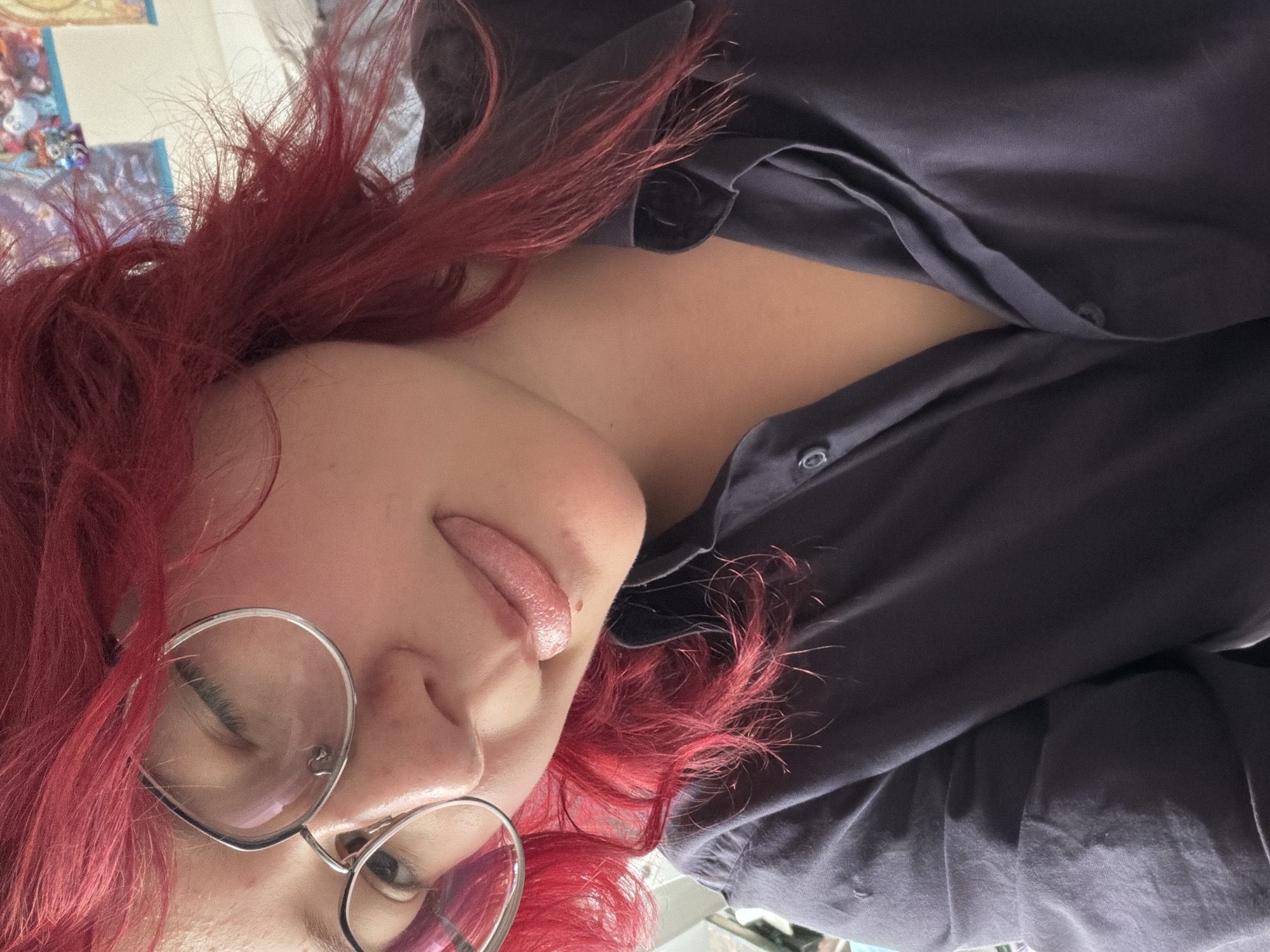 A close-up selfie shows a person with vibrant red hair wearing a dark collared shirt. The person is wearing heart-shaped glasses and is sticking their tongue out, winking. The background is blurred and appears to be an indoor setting with posters on the wall. There is no discernible information about the location in Chelyabinsk, Russia, as the image focuses solely on the subject.