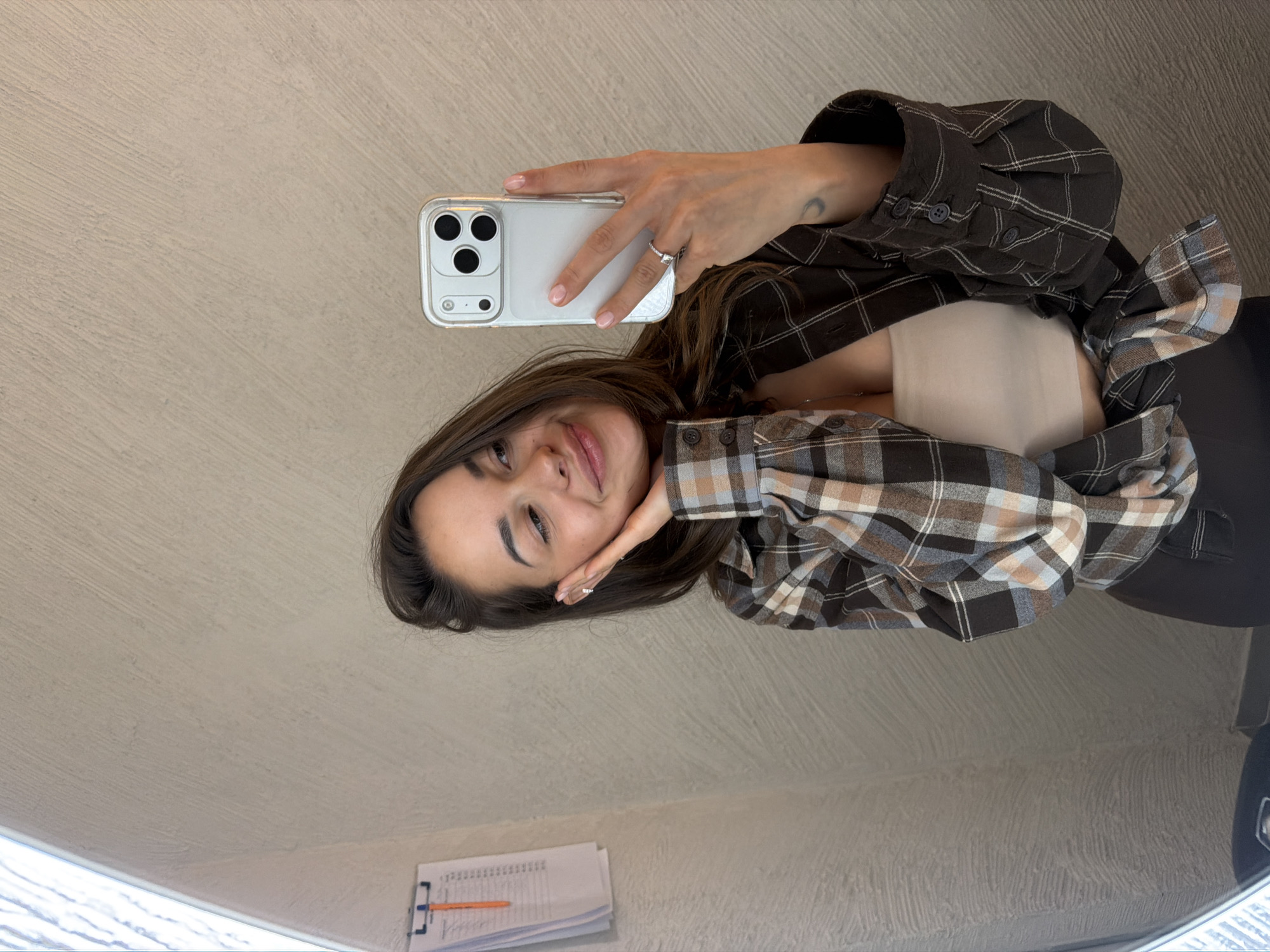 This mirror selfie, taken indoors, features a young woman in Krasnoyarsk, Russia. She is smiling gently at the camera, her head tilted slightly, with her long, dark brown hair cascading around her. She holds a silver smartphone with a clear case and a triple-lens camera in her left hand, while her right hand rests lightly near her chin.
The woman is wearing a brown and beige plaid flannel shirt, unbuttoned to reveal a light, possibly cream-colored, bandeau top or bra underneath. A silver-toned ring is visible on her left ring finger, and a small crescent moon tattoo is noticeable on her left forearm.
The background consists of a light beige, textured wall. In the lower reflection of the image, on a surface below the woman, several sheets of paper with handwritten notes or numerical data are visible, along with a pencil, suggesting a personal or study setting. Her expression conveys a calm and contented mood. The specific content of the visible text on the papers is not discernible, but it appears to be either writing or tabular data. The overall impression is a casual, relaxed moment captured indoors.