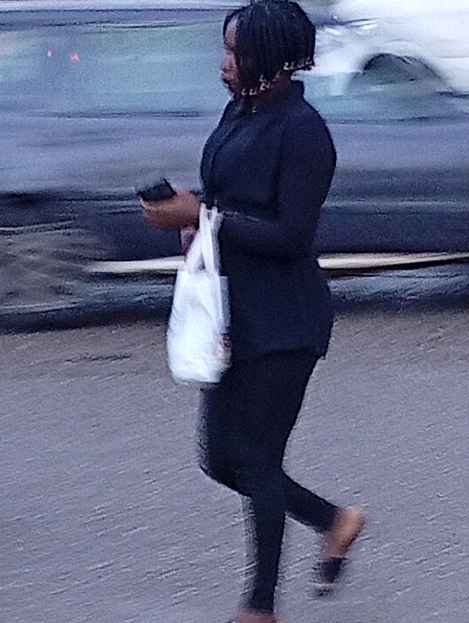 The image shows a full-body shot of a woman walking on a paved street, presumably in Jos, Nigeria. She is wearing a dark jacket over dark leggings and appears to be barefoot or wearing very thin footwear. In her left hand, she holds a white plastic bag, and in her right hand, she holds a dark rectangular object, possibly a phone. Her hair is dark and styled in short dreadlocks, with decorative earrings visible. The background consists of a dark vehicle with its lights on, suggesting it is either day or dusk. The street surface appears wet or reflective.