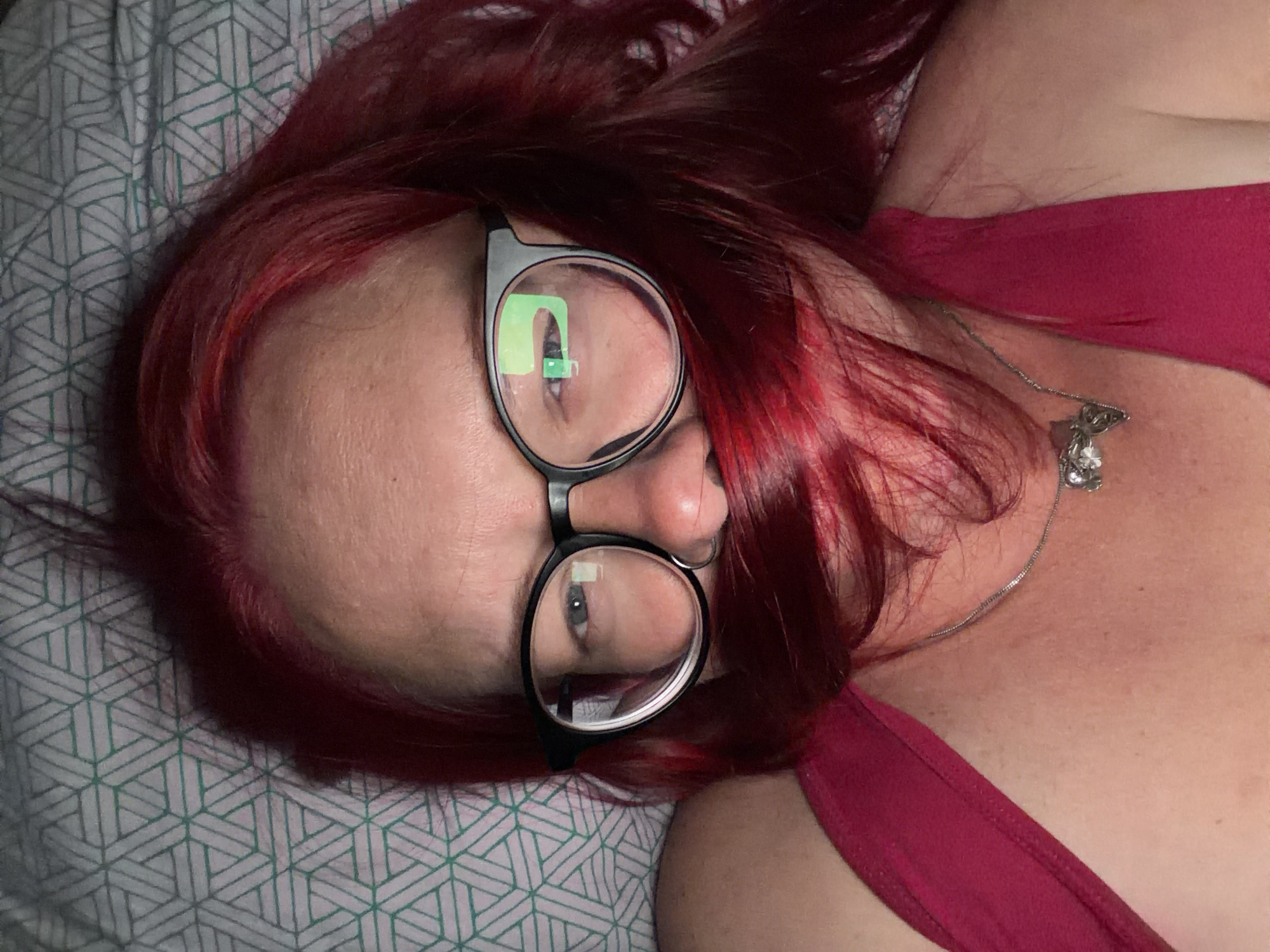 An image taken in Chaikovsky, Russia, shows a person with vibrant red hair, lying down and looking towards the camera. The subject is wearing dark, round-framed eyeglasses, through which their eyes are visible. A prominent, bright green reflection is seen in the upper lens of the glasses, suggesting a light source or screen directly in front. They have a subtle nose piercing.
The person is wearing a dark red or maroon sleeveless top, with a silver necklace adorned with a decorative, possibly floral, pendant visible on their chest and neck area. Their head rests on a pillow or bedding with a repeating geometric pattern in shades of teal or grey on a lighter background. The overall setting appears to be indoors, likely a bedroom or living space, given the presence of bedding. The lighting is soft, highlighting the texture of the hair and skin. No text is visible in the image.