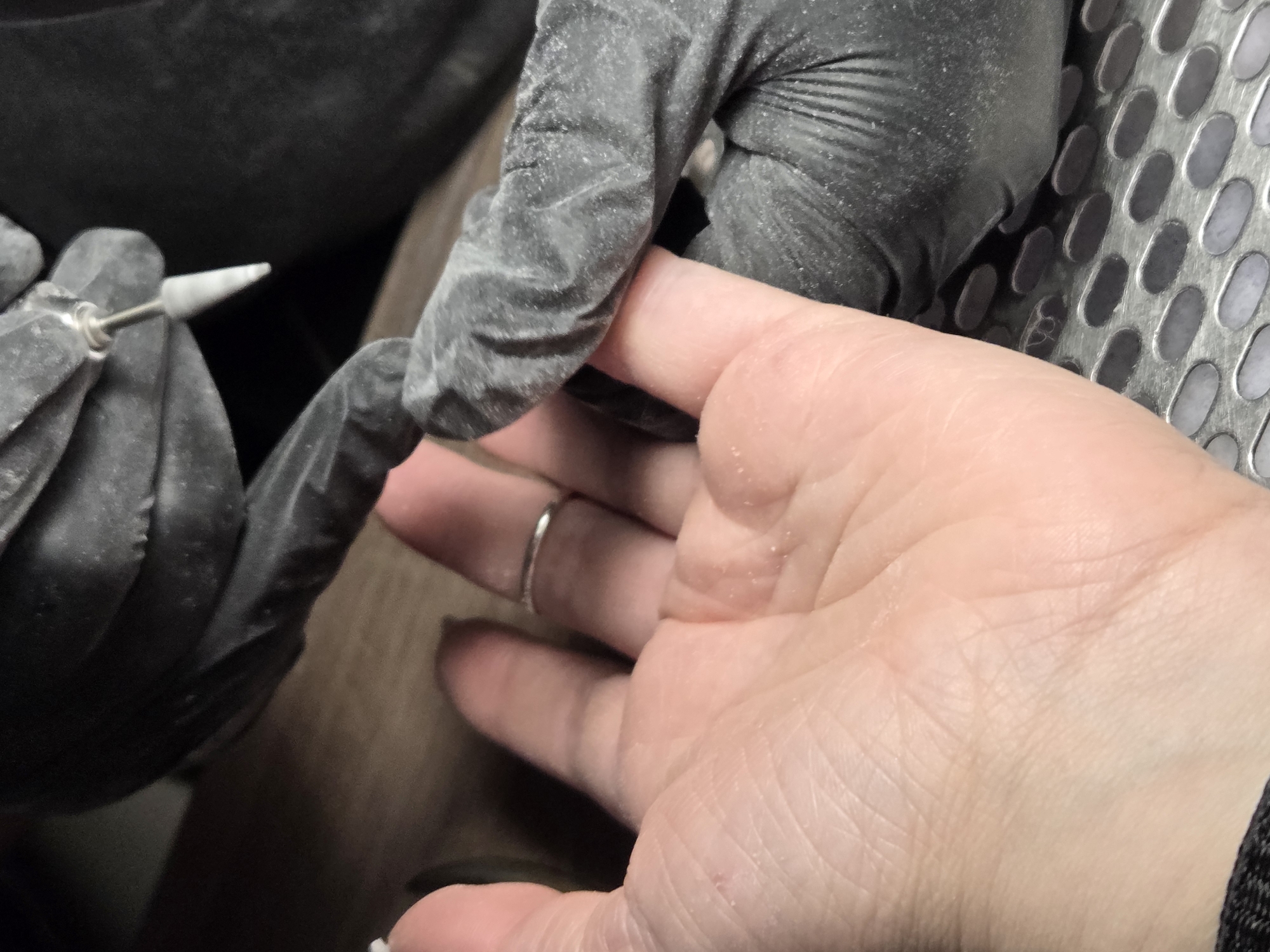 A gloved hand is holding a small rotary tool to work on a person's fingernail. The scene appears to be indoors with a metal grating in the background. The person's hand has a silver ring on one finger. The city and specific location are unknown.