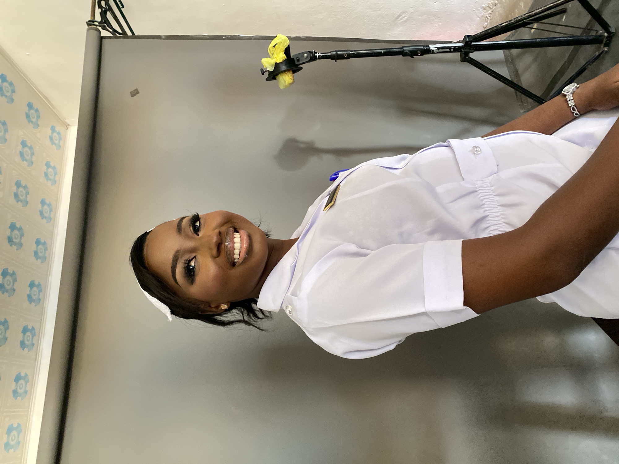 A medium shot depicts a smiling, dark-skinned woman wearing a white nurse's uniform and a nurse's cap. She has well-defined eyebrows and full makeup with bold false eyelashes. On her wrist, she wears an ornate silver watch. The background is a grey wall. A tripod for camera equipment is visible at the top of the frame with yellow cloth attached to the head of the light stand. The bottom of a wall with blue decorative tiling is barely visible on the left edge of the frame. It is not possible to determine the specific location in Gura Top, Nigeria.